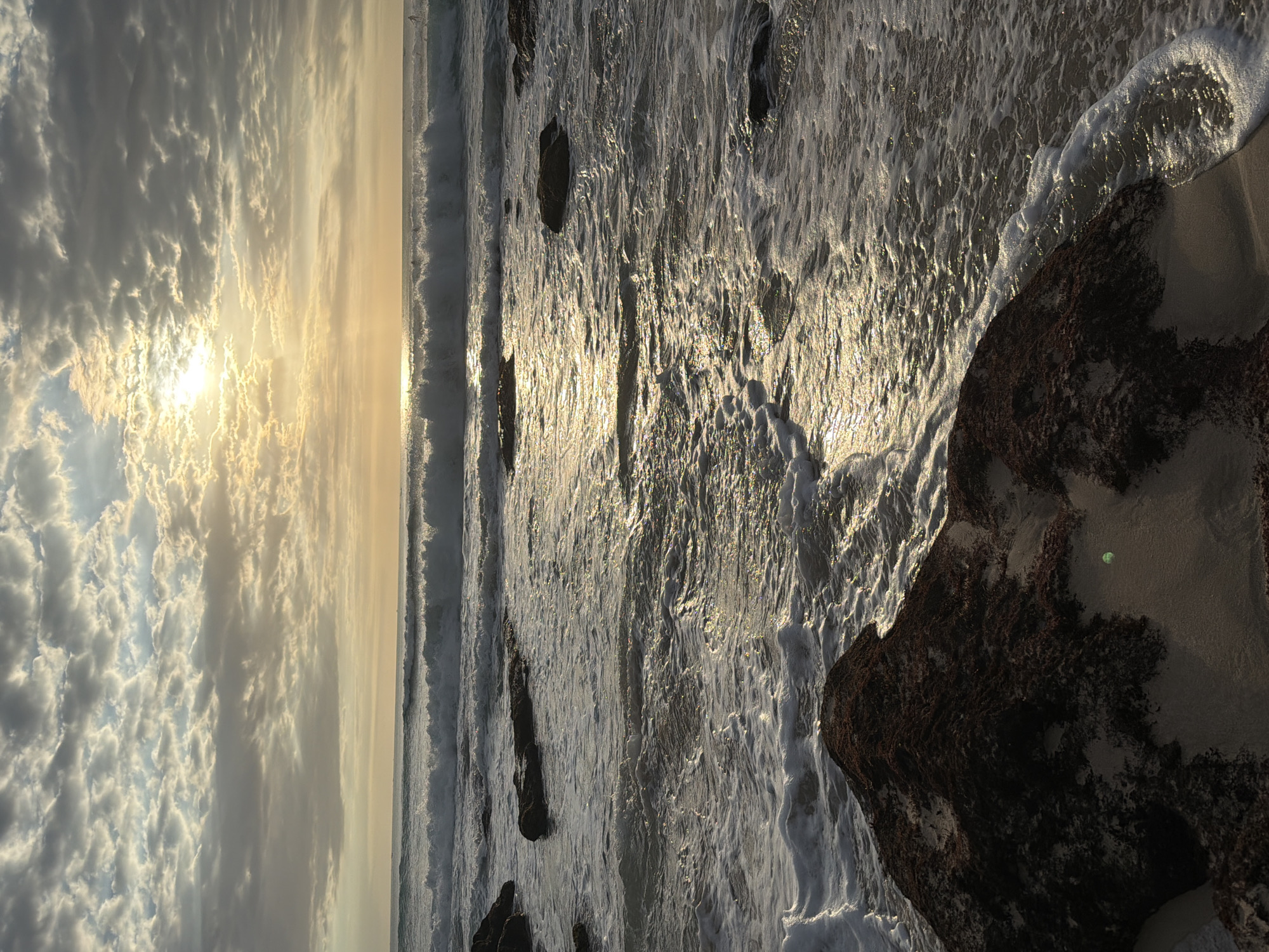 The image shows a coastal scene in Kuta Selatan, Indonesia, at sunrise or sunset. Waves are crashing onto a sandy beach with rocky formations. The sun is visible through a cloudy sky, casting a bright glow across the water. The waves are foamy and turbulent, reflecting the sunlight. A large, dark rock formation is present on the right side of the image, partially covered with sand. The beach appears deserted, with no people visible.