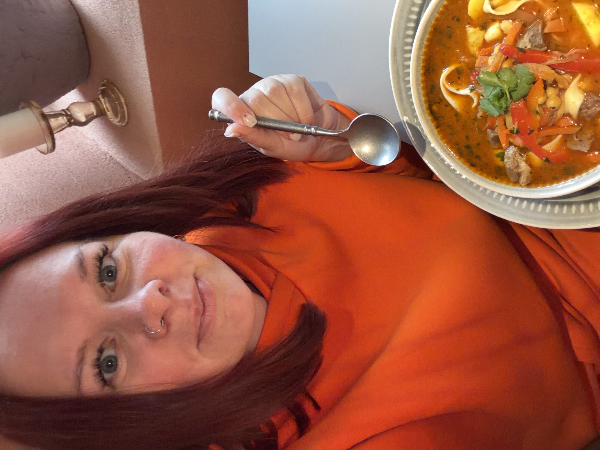 A selfie-style photograph features a woman with auburn hair, light skin, and blue eyes. She is wearing an orange top. She holds a silver spoon in her right hand, extended towards the upper right of the frame. A bowl of soup is visible in the upper right corner. The soup contains meat, vegetables, and noodles. The woman has a nose ring and visible eyelashes. In the background, there is a portion of a wall and a decorative object that may be part of a candle holder. There is no definitive information to confirm that the location is in Chaikovsky, Russia.