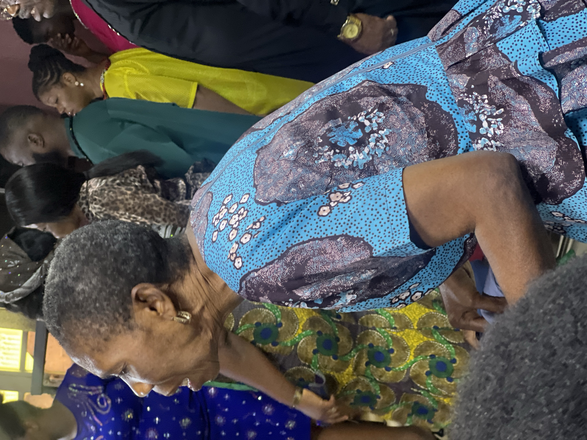 The image captures a close-up of a woman with short, grey hair, facing left with her mouth slightly open, as if speaking. She is wearing a vibrant blue dress adorned with a pattern of dark blue and white floral designs and small black dots. Her ear is visible with a hoop earring. Behind her, other individuals are partially visible, including a person in a bright yellow top and another in a green garment. The background suggests an indoor setting, possibly a gathering or event. The overall scene appears to be a candid moment captured during a social occasion.