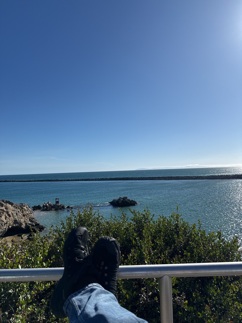 The image shows a view of the Pacific Ocean in Newport Beach, California, United States, on a clear day. In the foreground are a pair of feet wearing dark hiking shoes and blue jeans, resting on a silver railing. Behind the railing, there is dense green shrubbery. The middle ground features rocky formations and a small lifeguard tower in the water, leading out to a long breakwater or jetty extending into the ocean. The sky is a bright, clear blue, with the sun appearing at the top right. The ocean is calm with sunlight reflecting on the surface.