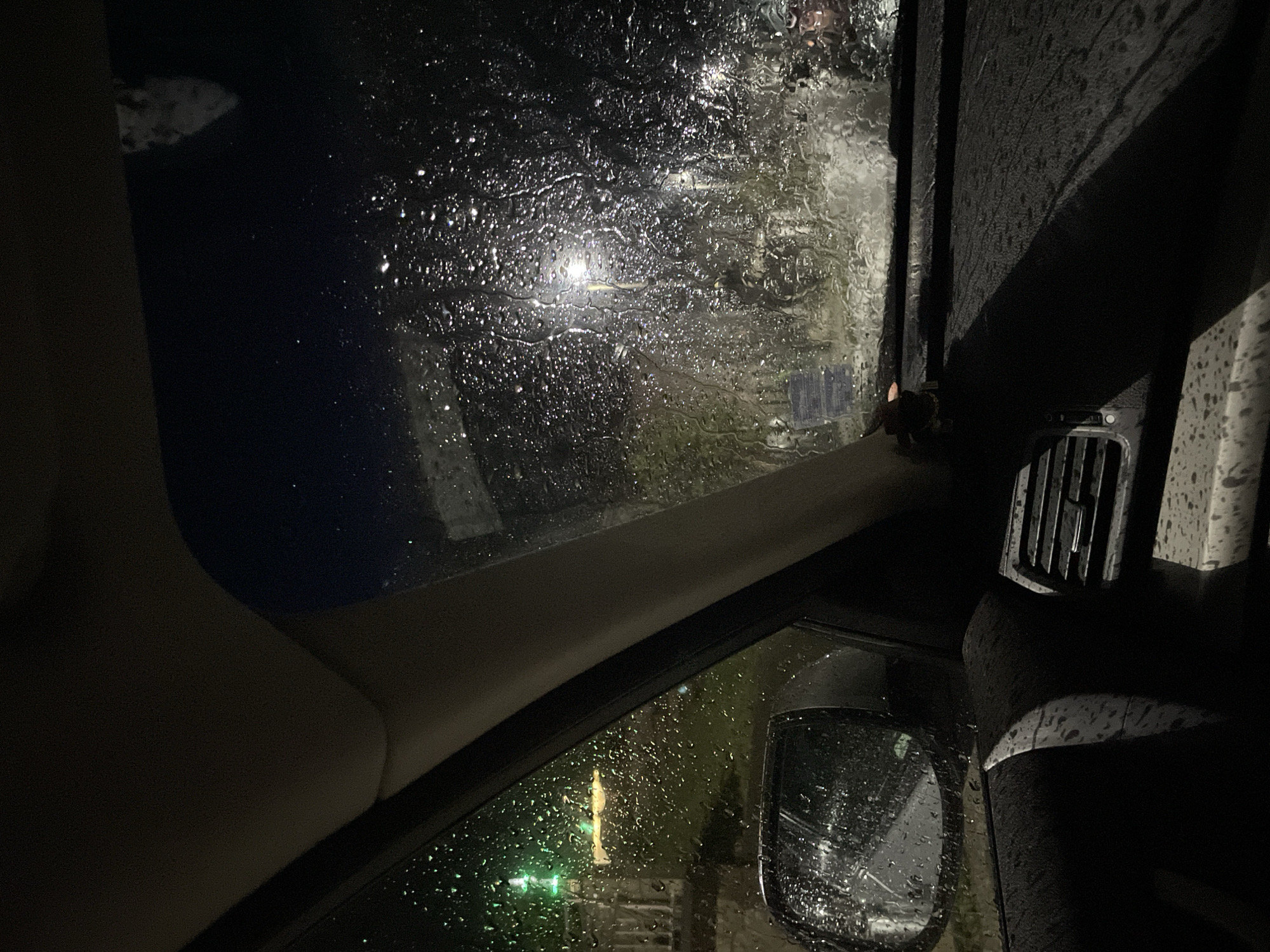 The media file captures a view from inside a car, looking out through rain-splattered windows at night. Water droplets and streaks heavily cover the windshield and side windows, blurring and distorting the exterior environment.
Through the wet glass, artificial light sources appear as bright, scattered blurs, including white streetlights and distinct green lights, suggesting an urban or suburban setting. The outdoor scene is dark, confirming nighttime and heavy rain.
Inside the vehicle, elements like a light-colored A-pillar on the left, a dark dashboard, and an air vent on the right are visible. A small, dark object, possibly an insect, clings to the exterior of the A-pillar near the window. The side mirror in the lower right is also heavily covered in raindrops. A yellow or gold-colored object, likely a hanging ornament, is suspended near the side window. Parts of the car's seating are visible in the foreground.
The image conveys the experience of being inside a vehicle during a rainy night in Startsevo, Bulgaria, focusing on the immediate car interior and the obscured, wet view outside. No legible text is visible.