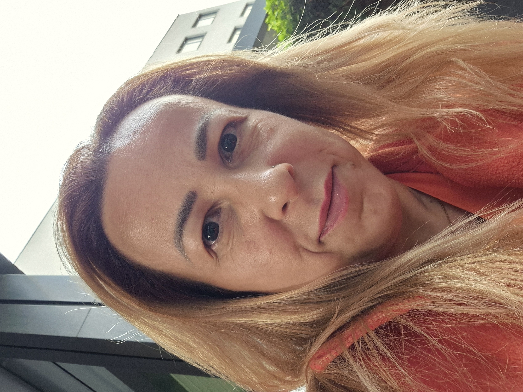 A medium shot selfie of a fair-skinned woman with light brown hair, taken outdoors. She has dark eyes, a small nose, and pink lips. She is wearing an orange hooded jacket. The background shows a modern building with windows and some greenery. The lighting suggests it is daytime, possibly with overcast skies. The camera is positioned slightly above and to the side of her face.