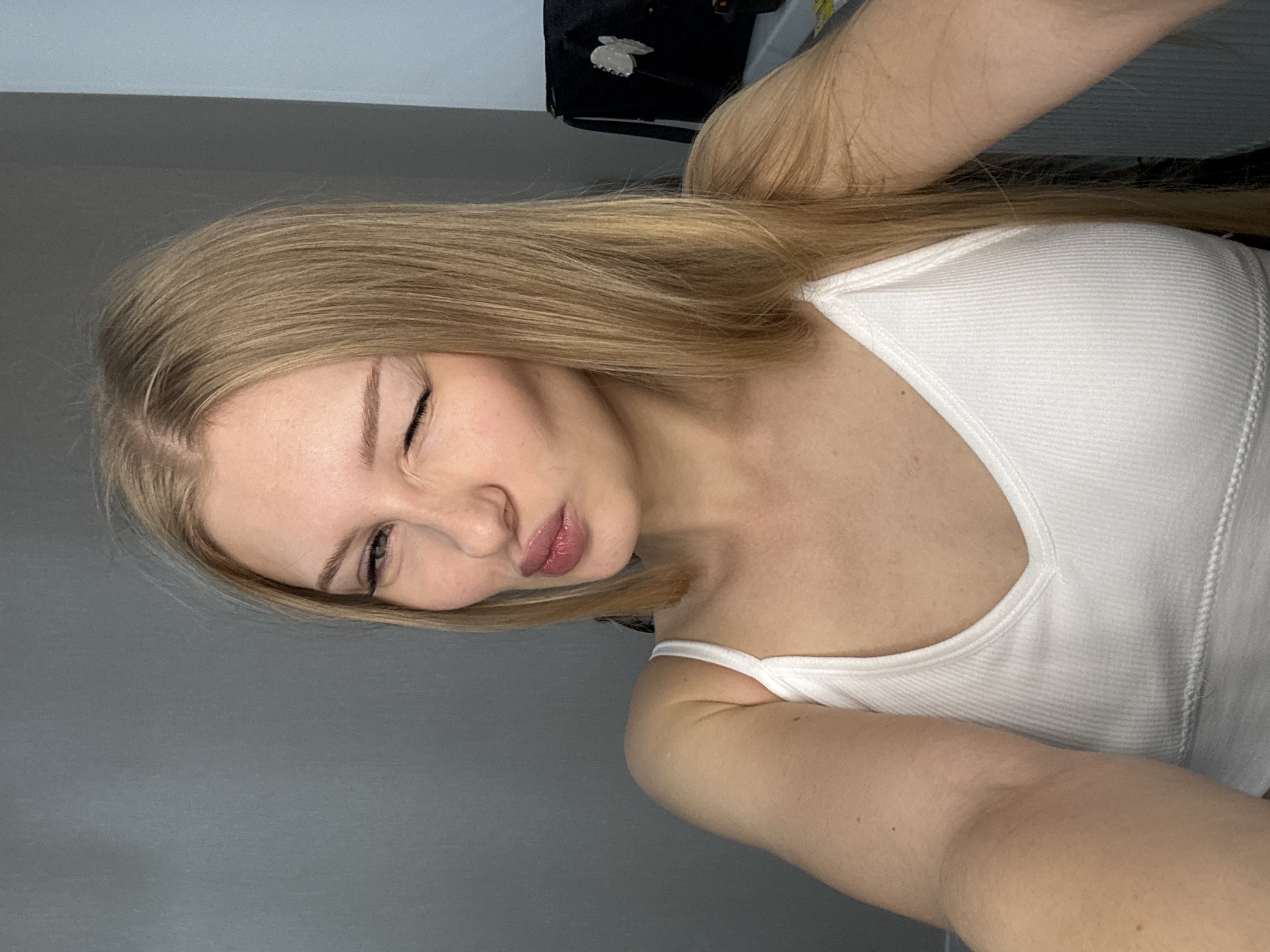 A young woman with blonde hair is taking a selfie in what appears to be a residential setting. She is wearing a white tank top. Her right eye is closed in a wink, and her lips are pursed. The background is a neutral gray wall. There are no identifiable landmarks or details to indicate the specific location within the city of Perm, Russia. The image is a close-up, focusing on the woman's face and upper body.