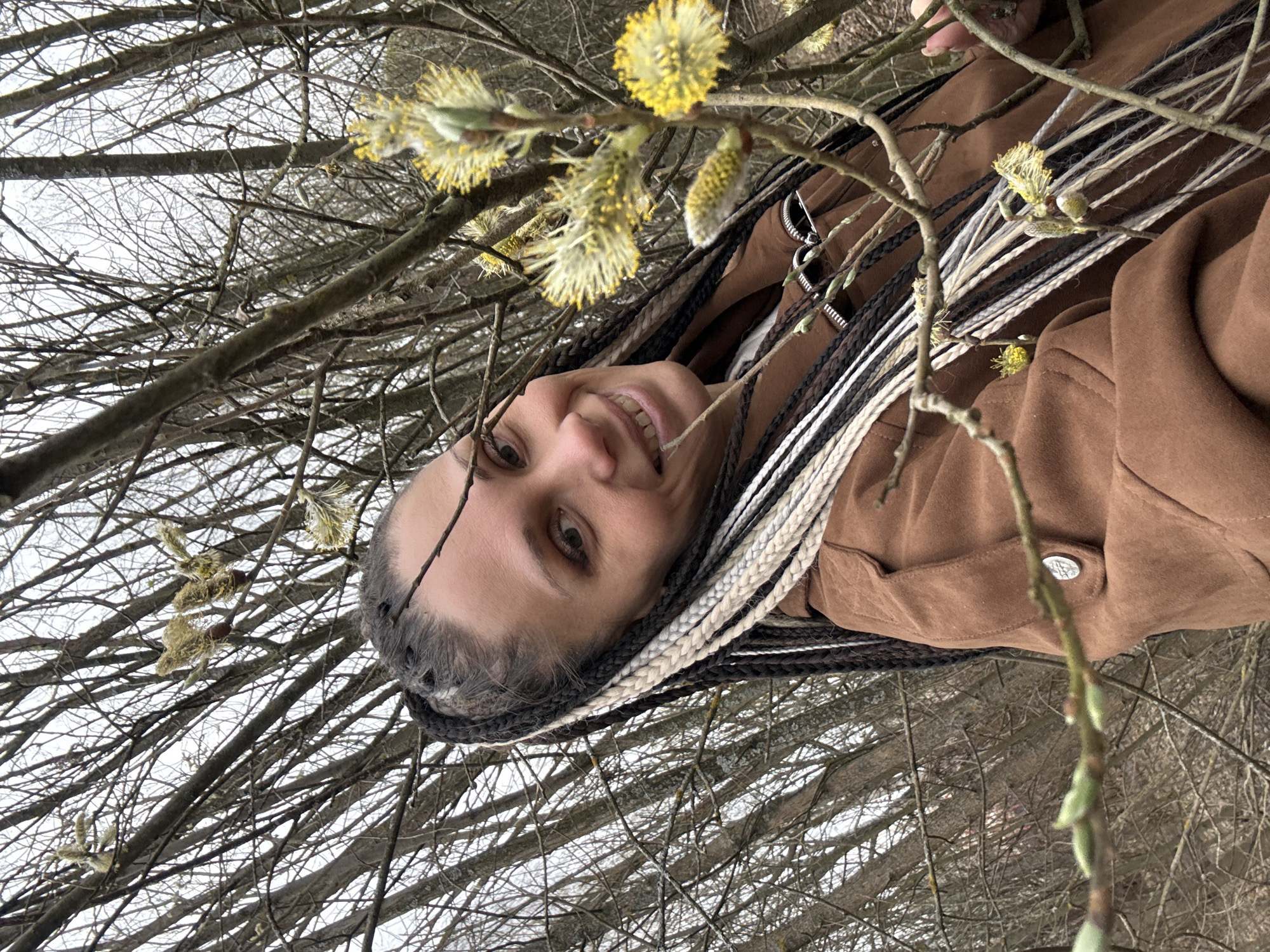 The image shows a woman with dark hair and multiple braids, some black, some white, smiling while standing among branches with emerging yellow flowers. She is wearing a brown jacket. The background is a sky seen through the branches of bare trees. There is no information to confirm that this image was taken in Rusanovo, Russia.