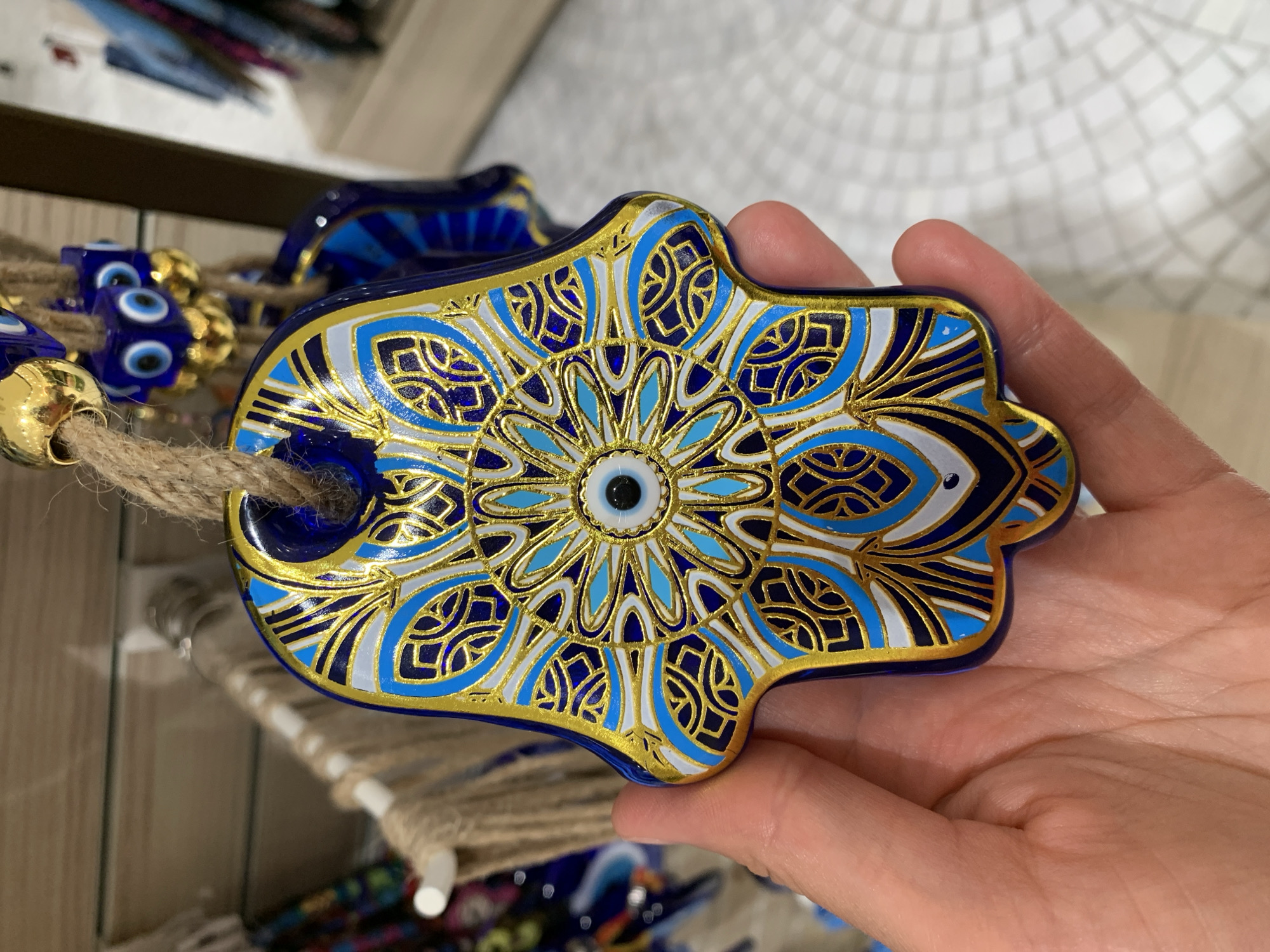 A hand holds a blue and gold Hamsa hand charm with an evil eye design. The charm is decorated with intricate patterns in shades of blue, white, and gold. It is suspended by a rough twine. In the background, out-of-focus, are shelves with various items, including what appear to be more blue glass charms. The flooring is visible at the bottom right and appears to be tiled. This image was taken in Aksu, Turkey.