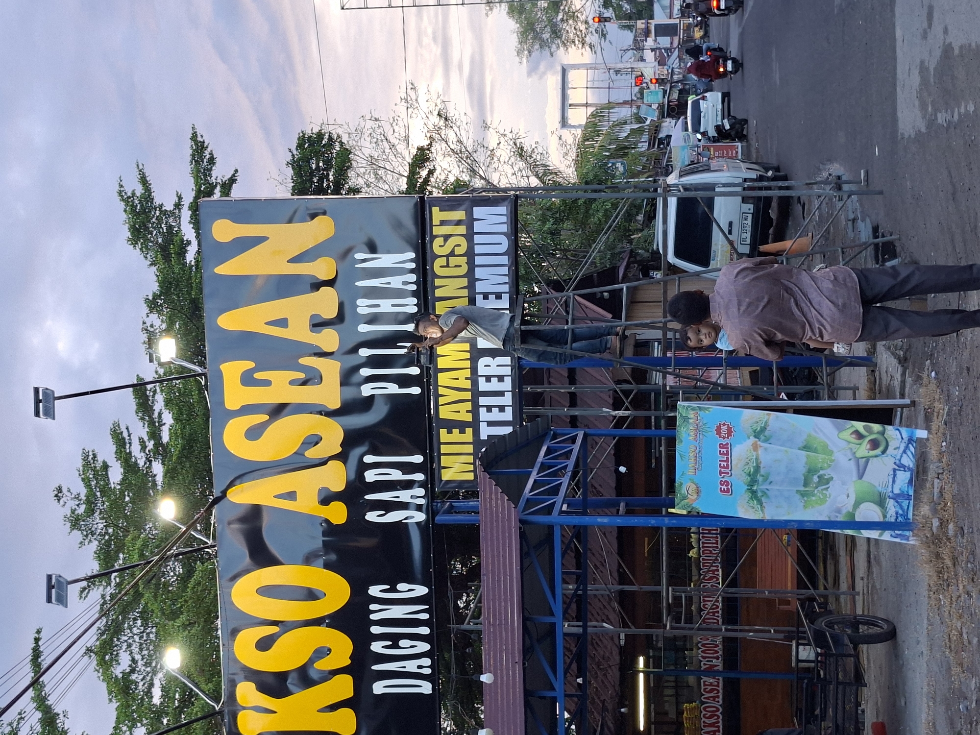 The image depicts an outdoor scene in Lhokseumawe, Indonesia. A large black and yellow sign displays the words "Bakso Asean," and under that "Daging Sapi Pilihan, Mie Ayam Pangsit Teler Kemium." Two men work on scaffolding in front of a street with light traffic of cars and motorcycles. An advertising poster for "Es Teler 201" is partially visible. A man holds a child. Street lighting is visible and the sky is overcast. Vehicle registration "BL 1292 NV" is visible on a white car.