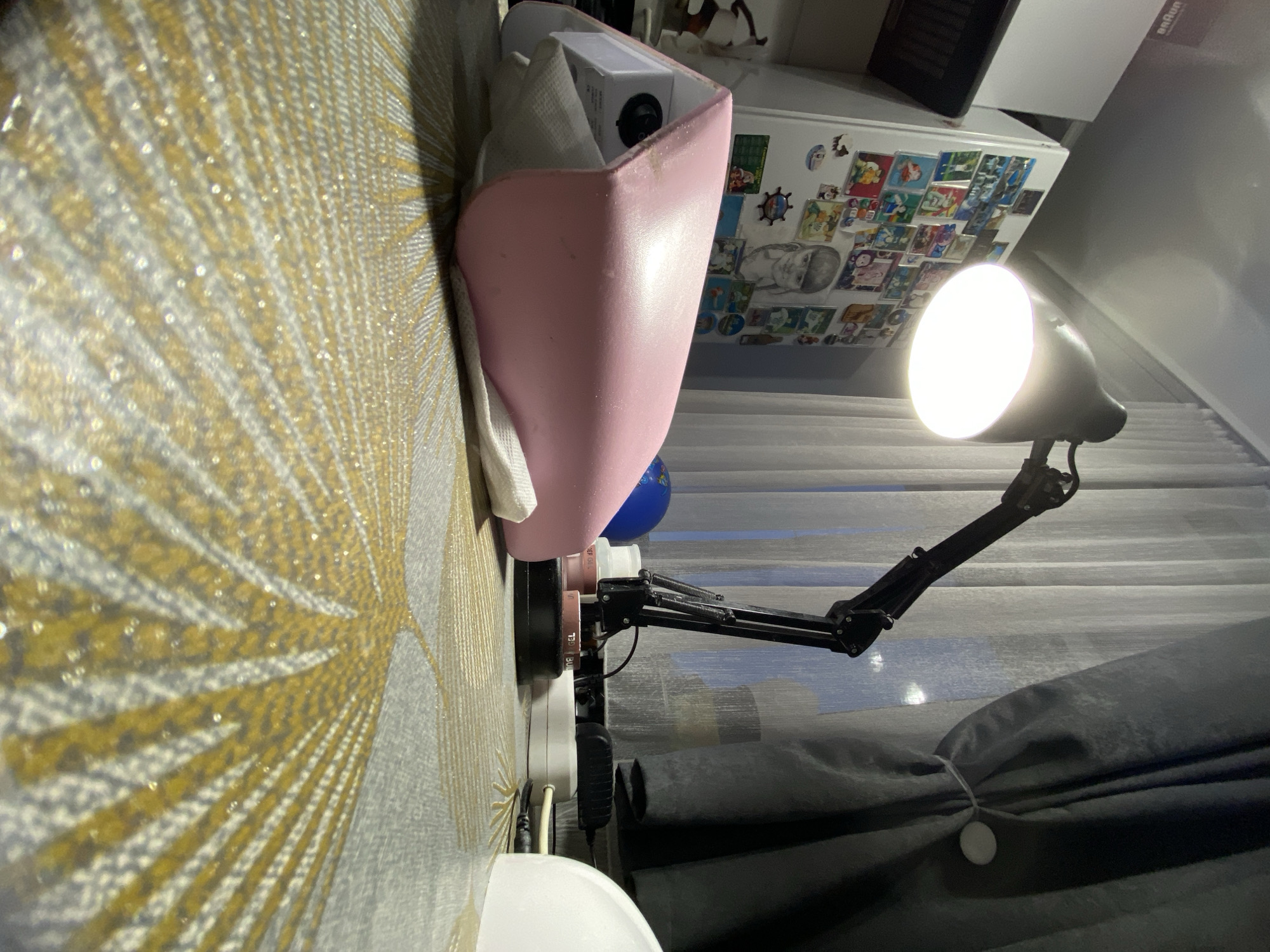The image depicts a dimly lit indoor scene, possibly a home office or crafting area, featuring a desk with various items. A prominent desk lamp with a bright LED bulb is positioned to the right, illuminating the workspace. In the foreground, a pink object with a white cloth underneath is placed on a patterned surface, likely a tablecloth or desk mat. To the left, a refrigerator is visible, adorned with numerous magnets and possibly pictures. Behind the desk lamp, sheer curtains allow some natural light to filter through, and a darker curtain is drawn to the side. The overall ambiance is one of a personal space, likely in an apartment or house. The location is not specified within the image itself, and therefore, it is impossible to determine its specific location within the city of Ufa, Russia, or any other city.
