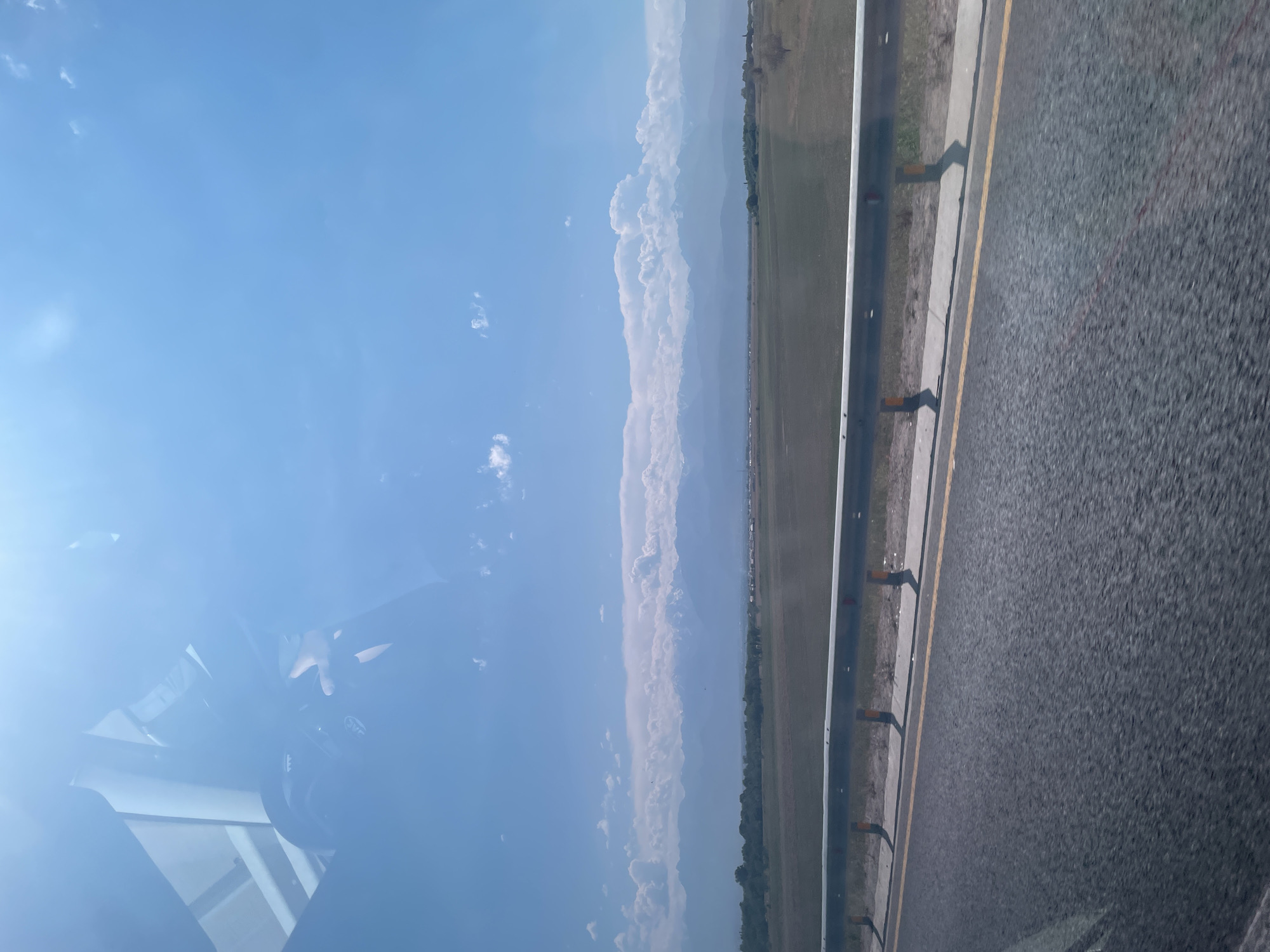 The image captures a wide-angle view of a landscape from a moving vehicle, likely on a highway. In the foreground, the asphalt road surface with a yellow dividing line is visible, suggesting a paved route. A metal guardrail with yellow and red reflectors runs along the side of the road. Beyond the guardrail, a flat, grassy plain stretches towards a distant mountain range. The mountains, capped with snow, are shrouded in a hazy blue due to atmospheric perspective. The sky above is a clear blue with scattered white clouds, indicating fair weather. Reflections are visible on what appears to be the vehicle's window, obscuring a portion of the upper left corner of the image and showing a partial view of a helmet and a hand, implying the presence of passengers. The scene is likely set in the vicinity of Baiserke, Kazakhstan, given the characteristic landscape featuring plains and distant snow-capped mountains.