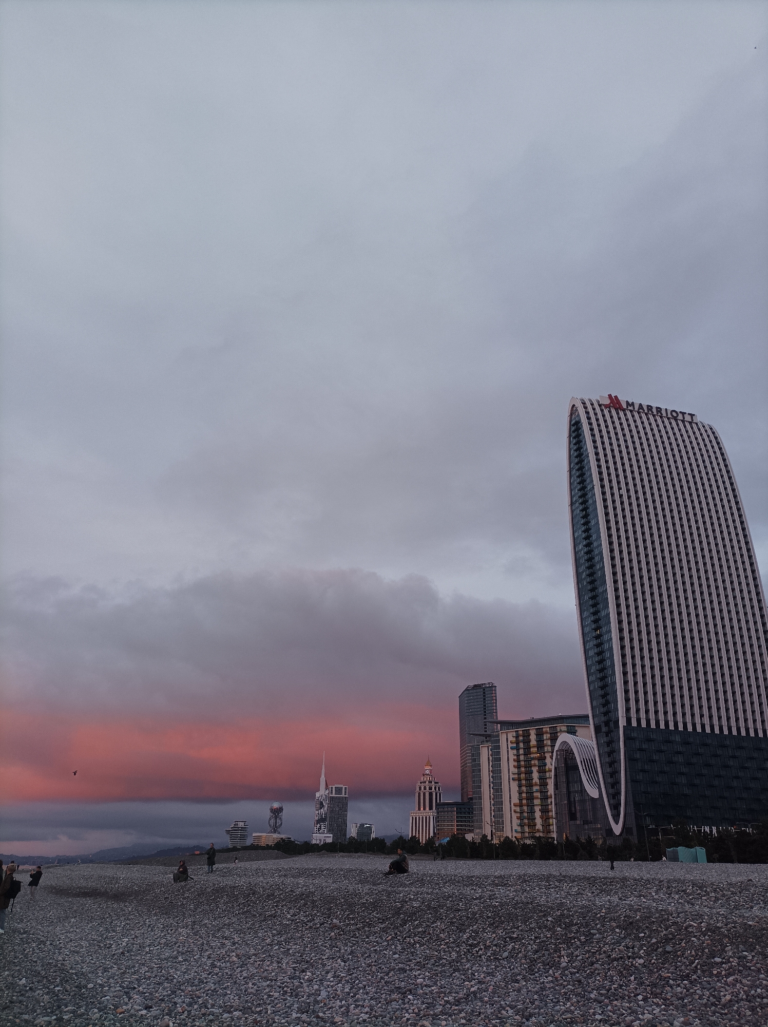 The image presents a dusk scene on a pebble beach in Batumi, Georgia. The foreground is composed of a dark grey pebble beach, where approximately eight to ten indistinct human figures are sparsely distributed, mostly standing or seated, without clear active interactions.
The midground and background showcase the modern urban skyline of Batumi. Dominating the right side is the Le Méridien Batumi (formerly Batumi Tower), a tall, curved skyscraper with a light-colored, ridged facade and "MARRIOTT" branding at its summit. To its left, a series of other contemporary high-rise buildings are visible, including one with a distinctive arched architectural feature. Further to the left, the Batumi Alphabet Tower, with its spherical top, and portions of the Batumi Ferris Wheel are identifiable landmarks.
The sky is largely overcast, with dense, dark grey clouds covering the upper two-thirds of the frame. A prominent horizontal band of vibrant pink and orange clouds stretches across the horizon, suggesting twilight conditions. Below this colorful band, a dark, silhouetted landmass, likely distant mountains or hills, is discernible. A single dark bird is captured in flight against the pink portion of the sky on the left.