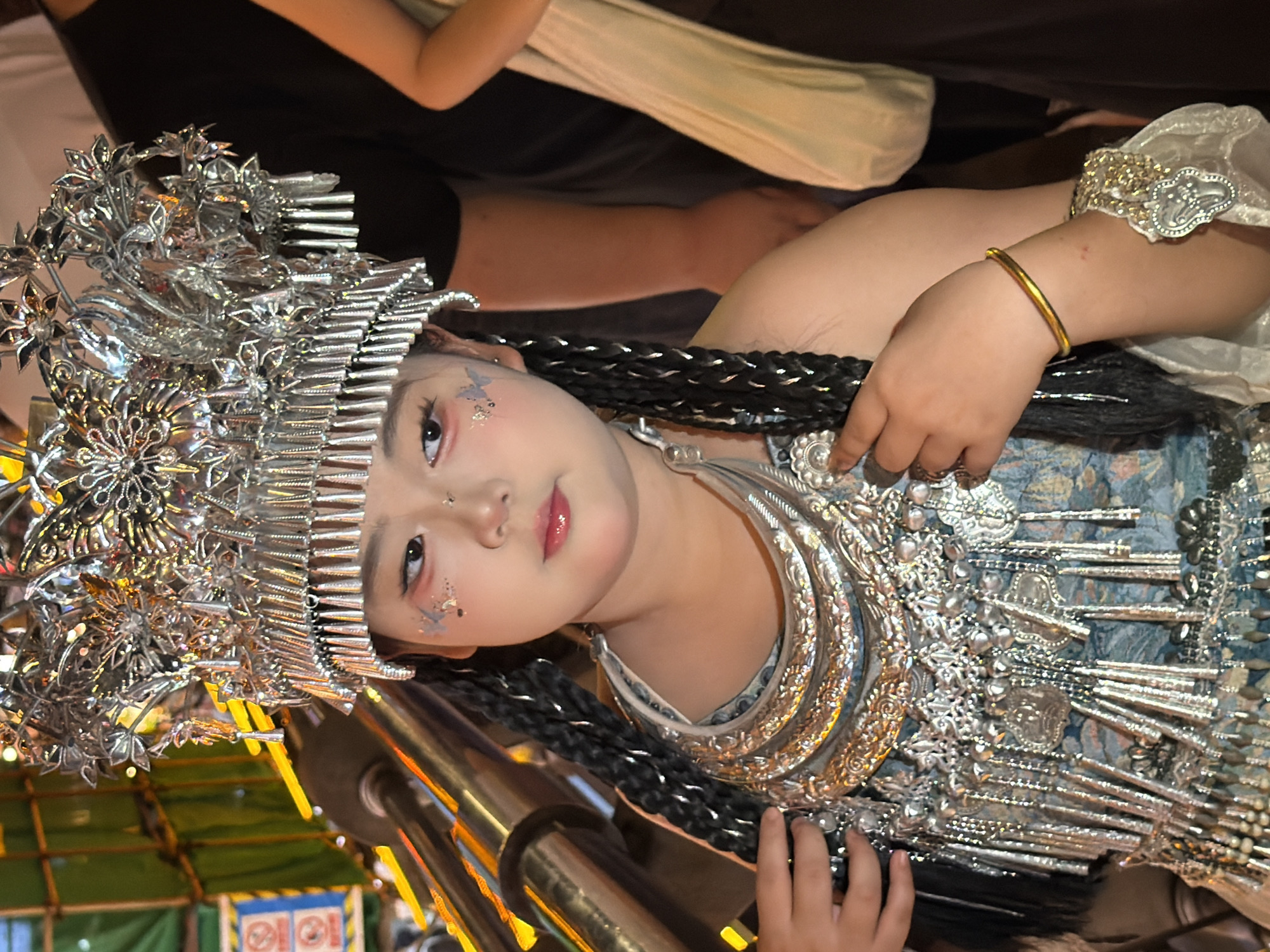 A young girl, adorned in traditional Chinese ethnic attire, is the central subject of this image. She wears an elaborate silver headdress, intricately designed with floral and geometric patterns. Her costume is equally ornate, featuring a blue and floral patterned garment embellished with numerous silver ornaments and dangling chains. She has distinctive makeup, including glitter on her cheeks and styled eyebrows. Her braided black hair is partially visible. The girl wears a gold bangle on her left wrist, and her right hand, with rings on several fingers, rests on her chest.
The scene appears to be indoors or under some form of canopy, with a glimpse of what looks like a market stall or an outdoor event in the background, indicated by green tarps and a visible sign with international prohibition symbols. The lighting suggests an artificial setting, possibly in the evening or a dimly lit interior.
The girl's pose and the close-up framing emphasize her regal and somewhat solemn expression, with a slight tilt of her head and her gaze directed away from the viewer. The overall impression is one of cultural celebration or performance, possibly a festival or a traditional event.
The image's orientation is rotated, with the subject appearing sideways. The location context of Yuzhong District, China, suggests the cultural elements are likely representative of a Chinese ethnic minority group. No discernible text is clearly legible in the image.