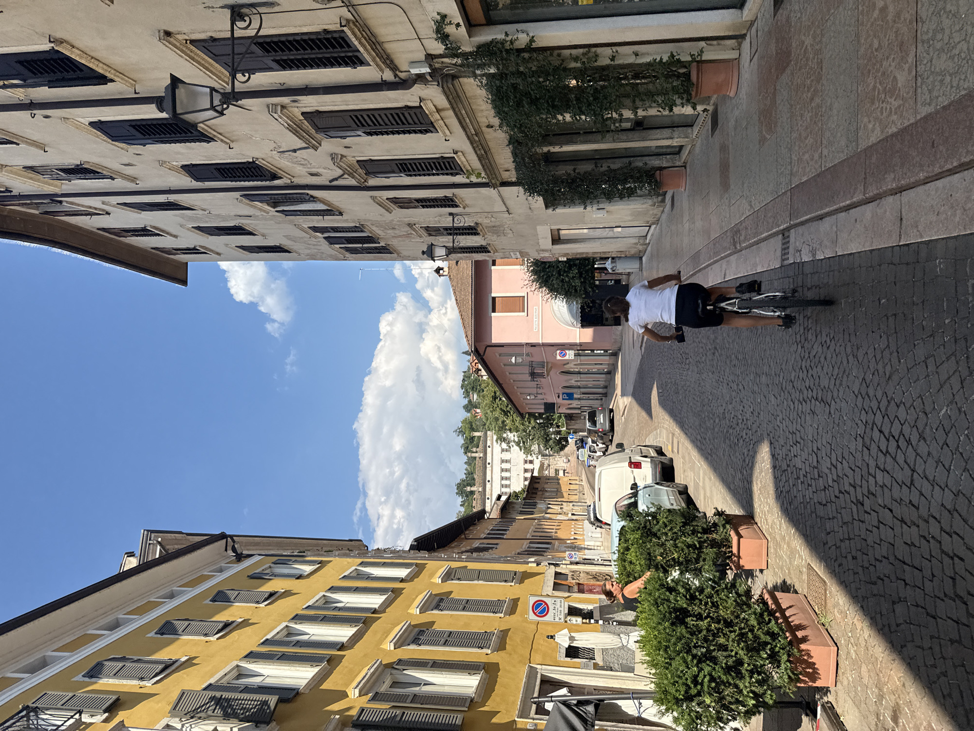 The image depicts a street scene in Trento, Italy, with a woman cycling away from the camera down a cobblestone street. She is wearing a white t-shirt and black shorts. The street is lined with buildings of various colors, including a prominent yellow building on the left with closed shutters and a pale beige building on the right also with shutters and a street lamp. Green potted plants are placed along the sidewalk. In the background, there are more buildings, some with red roofs, and a large white building with classical architecture is visible further up the hill, surrounded by trees. The sky is blue with scattered white clouds. There are parked vehicles on the right side of the street. The overall atmosphere is that of a sunny day in a European city.