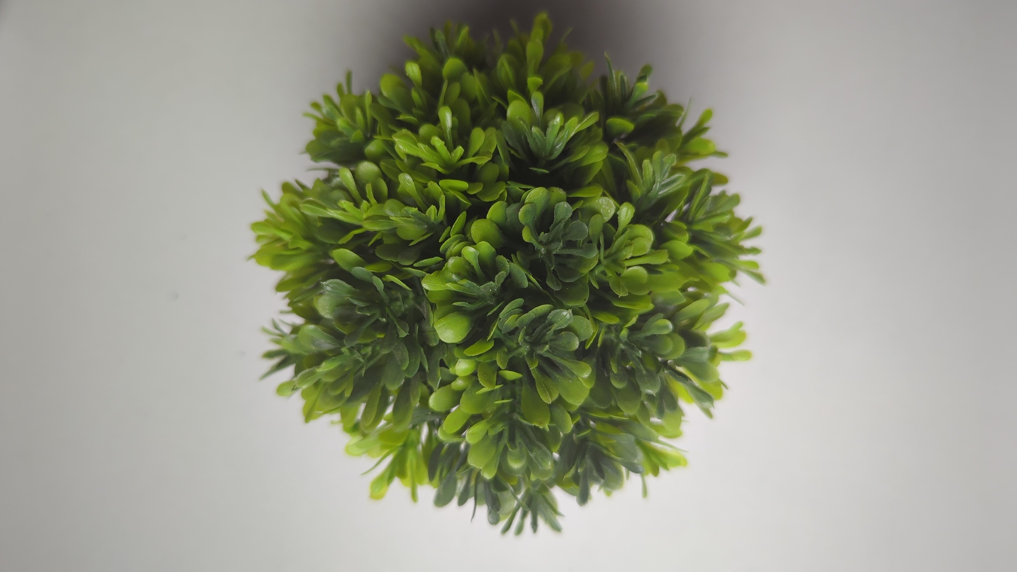 A top-down, close-up shot shows a spherical artificial plant with dense, bright green leaves against a plain, light gray background. The plant is centered in the frame, casting a subtle shadow that blends into the background. No other objects or people are visible in the scene. The image does not contain any elements that would indicate its location in Penjor, Indonesia.