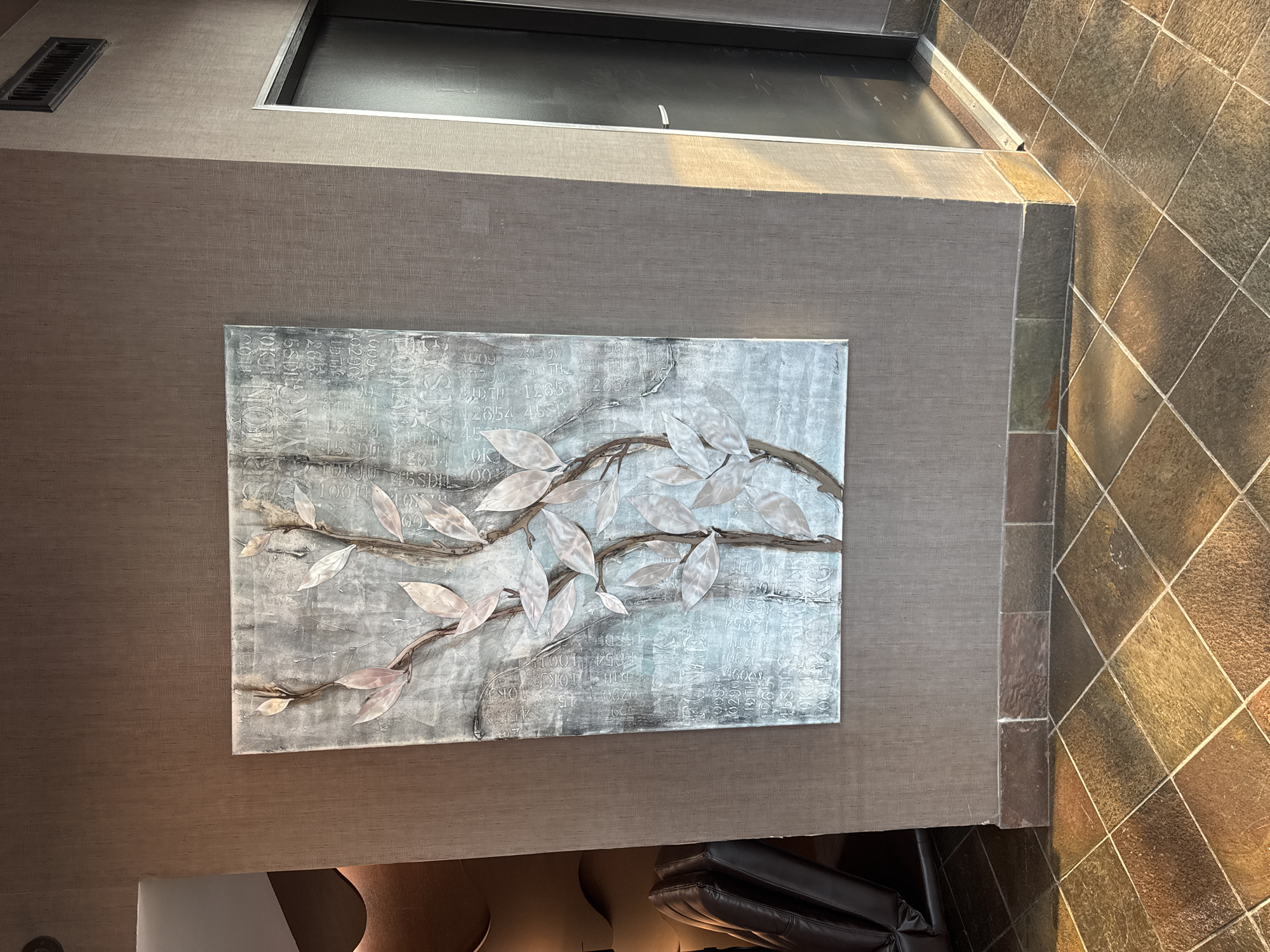 The image depicts a textured wall with a piece of art featuring a stylized tree branch with pale leaves. The artwork is hung on the wall, which has a muted brown texture. Below the artwork is a decorative border made of stone tiles, and the floor is tiled with large square tiles in various shades of brown and gold. To the right of the artwork is a dark door, and the wall adjacent to it shows a clear distinction between the shadowed and sunlit sides. In the background to the left, a portion of a dark leather chair and some curved, sculpted wall elements are visible. The lighting suggests it may be late afternoon, with sunlight casting distinct shadows on the floor and wall. The setting appears to be an indoor commercial or public space, possibly a hotel lobby or a reception area. The provided location information "North Vancouver, Canada" does not offer specific visual details to be incorporated into the description of the scene itself.