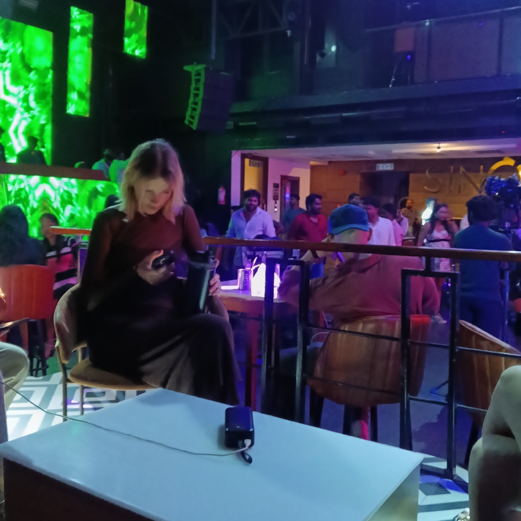 The image shows a dimly lit interior scene, possibly a nightclub or lounge, in Sinquerim, India. In the foreground, a light-colored rectangular table is visible with a black portable electronic device and a connecting cord on its surface. A woman with blonde hair, wearing a long brown dress, is seated at the table, looking at a mobile device in her hands. To her right, another person wearing a brown shirt and a blue cap sits on a chair. A black metal railing is situated between the foreground and the background, separating the seating area from the rest of the establishment.
In the background, there are several people standing and walking around. Some individuals are behind a bar or counter. The background is illuminated with a mix of colors, including blue, purple, and green lighting. A green-lit screen can be seen on the left side of the frame, displaying abstract patterns. The word "SIN" is partially visible on a wall in the background. The floor has a black and white geometric pattern. Speakers can be seen at the top of the frame.