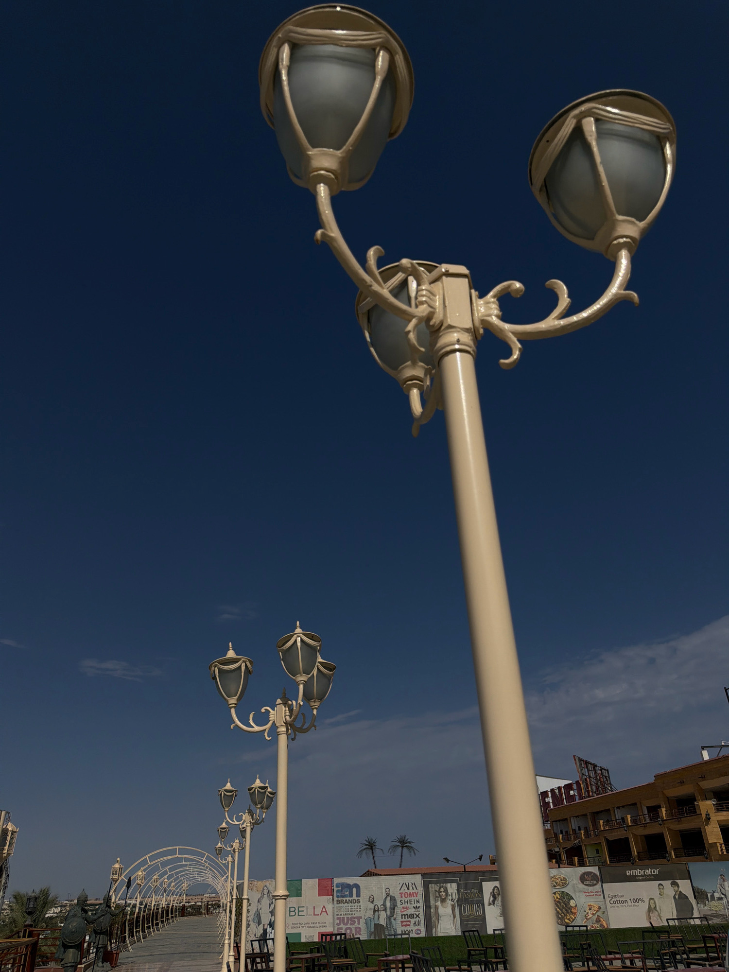 The photograph captures an outdoor scene in Sharm El Sheikh, Egypt, dominated by a tall, ornate street lamp with three globes, set against a clear, dark blue sky with a few wisps of clouds. In the mid-ground, a series of similar, but smaller, street lamps are visible, lining a walkway. The walkway itself is enclosed by a white, arched metal structure, reminiscent of a tunnel or canopy. To the left of the walkway, two bronze statues of armored figures holding shields and spears stand guard. Further to the right, a building with multiple stories, featuring a yellowish-brown facade and numerous windows, is visible. In front of this building, a large billboard displays various advertisements, including brands like "ZARA," "SHEIN," and "Cotton 100%." The ground in the foreground appears to be a paved area with several outdoor tables and chairs, suggesting a seating area for a cafe or restaurant. Palm trees are subtly visible in the background. The lighting indicates it is daytime, with bright sunlight casting shadows.