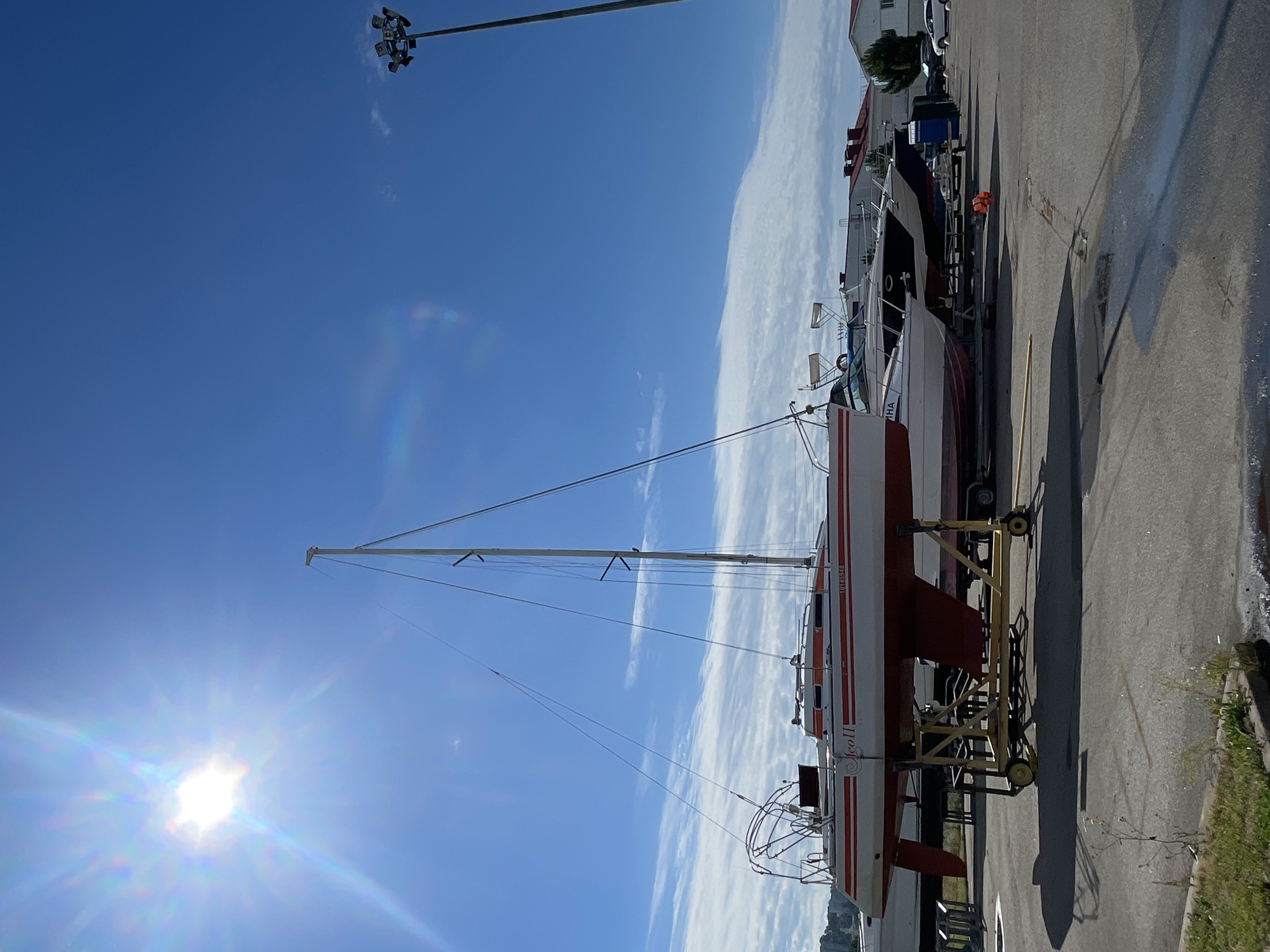 The image depicts a bright, sunny day with a clear blue sky, featuring a prominent sun with a lens flare in the upper left quadrant. Several boats are parked on land, some on trailers. In the foreground, a red and white sailboat is elevated on a yellow trailer. Behind it, other boats, including a dark-hulled motorboat, are lined up. In the background, buildings with red roofs and trees are visible. A tall light pole with multiple lamps stands to the right. The scene appears to be a boatyard or marina. The specific location within the city of Sochi, Russia, cannot be definitively determined from the visual information provided in the image alone.