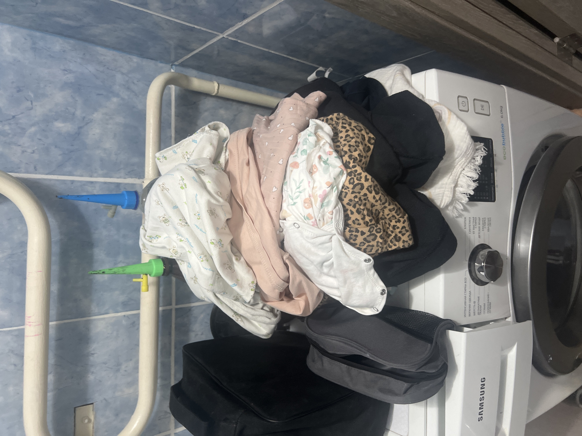 The image shows a pile of laundry on top of a Samsung washing machine in what appears to be a bathroom. The washing machine has a dial with various settings, including "ecoBUBBLE 6.0kg". There is also a black bag and a grey bag next to the laundry. The walls are tiled with blue and white tiles. In the background, there is a white pipe with pink marks on it, and two plastic cones, one green and one blue, attached to bottles. There are no people visible in the image. The scene suggests a domestic setting in Surgut, Russia, likely a laundry room or bathroom where laundry is being prepared for washing.