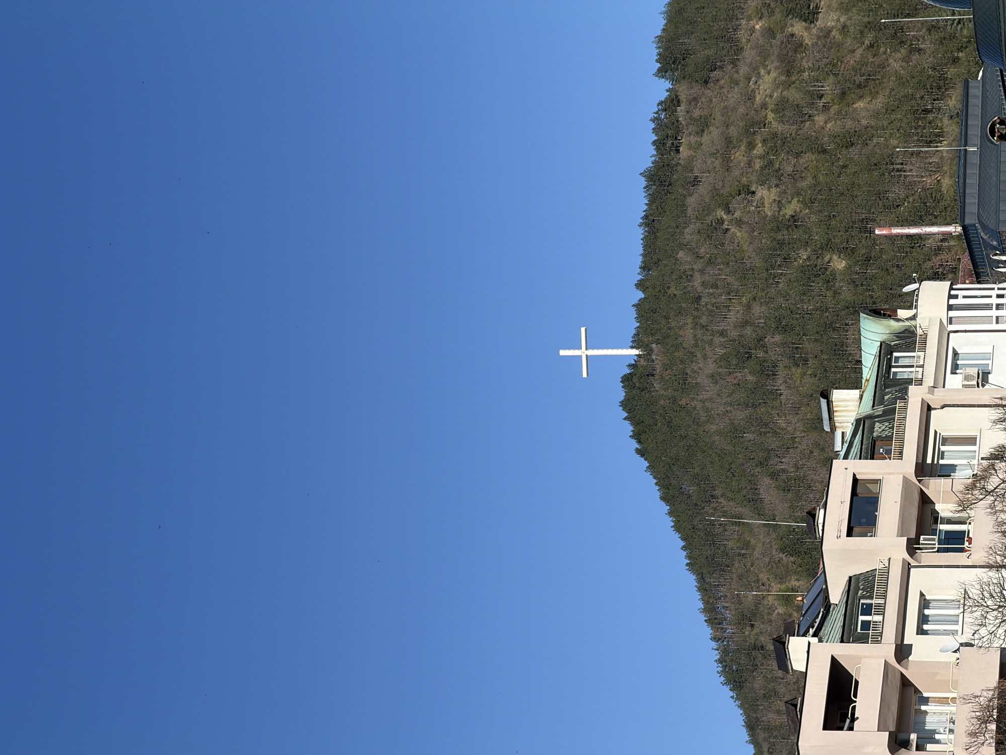 The image depicts a view of a white cross situated on a mountainside, with a clear blue sky occupying a significant portion of the frame. A dense forest covers the mountain. Adjacent to the mountain, a series of modern buildings, likely residential or commercial, can be seen. The architecture of these buildings is somewhat minimalist, featuring balconies and various windows. The cross and forest are landmarks in Blagoevgrad, Bulgaria.