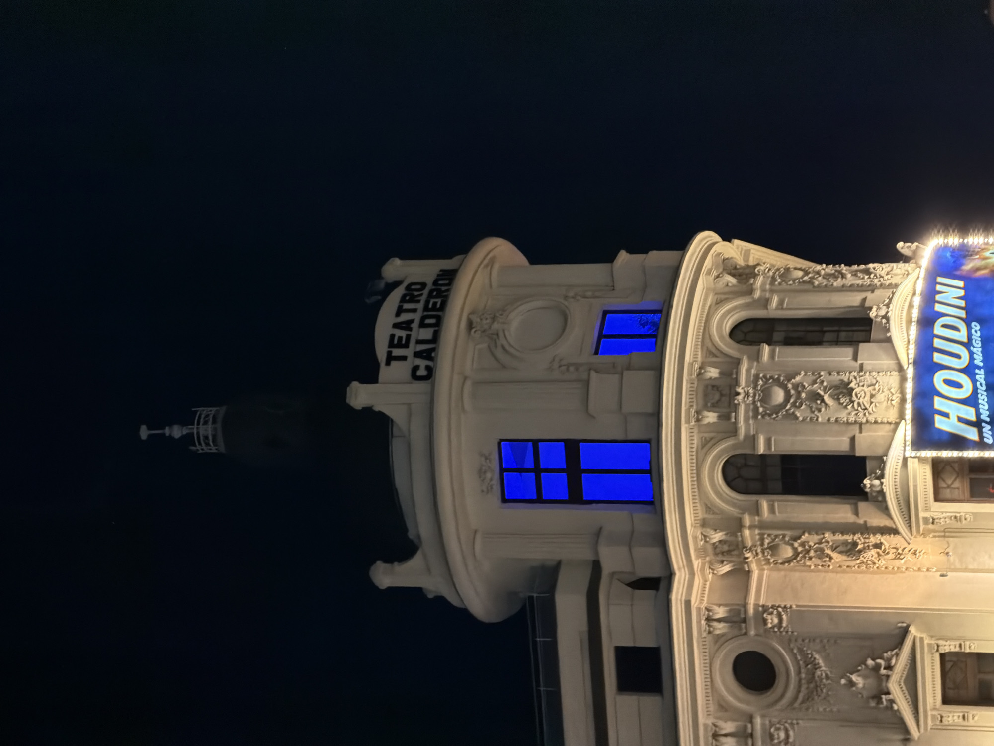 The image shows the Teatro Calderón in Madrid, Spain, at night. The building is illuminated, and a sign for the show "Houdini" is visible. The building's architecture is ornate, with decorative elements. The windows are lit with blue light. A spire is visible on top of the building to the left.
