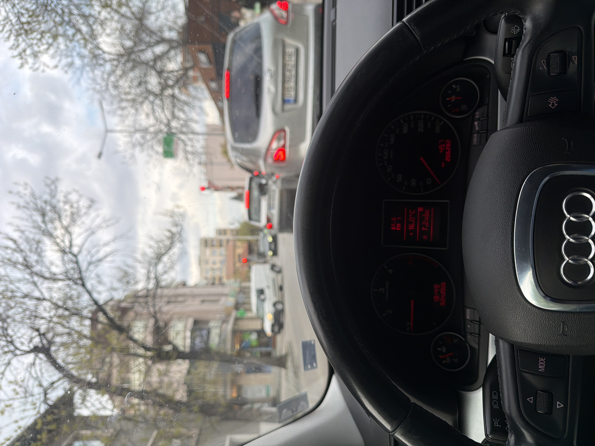 The image shows the perspective from the driver's seat of an Audi car. The steering wheel and dashboard are prominent in the foreground. The dashboard indicates a temperature of 16.0°C. Through the windshield, the image captures a street scene with cars, buildings, and trees. A silver car directly ahead has a license plate partially obscured but recognizable as "PB 7642HE," indicative of Bulgarian registration. The overall scene is consistent with a traffic environment in an urban area. Considering the license plate format, the photo was likely taken in Plovdiv, Bulgaria.