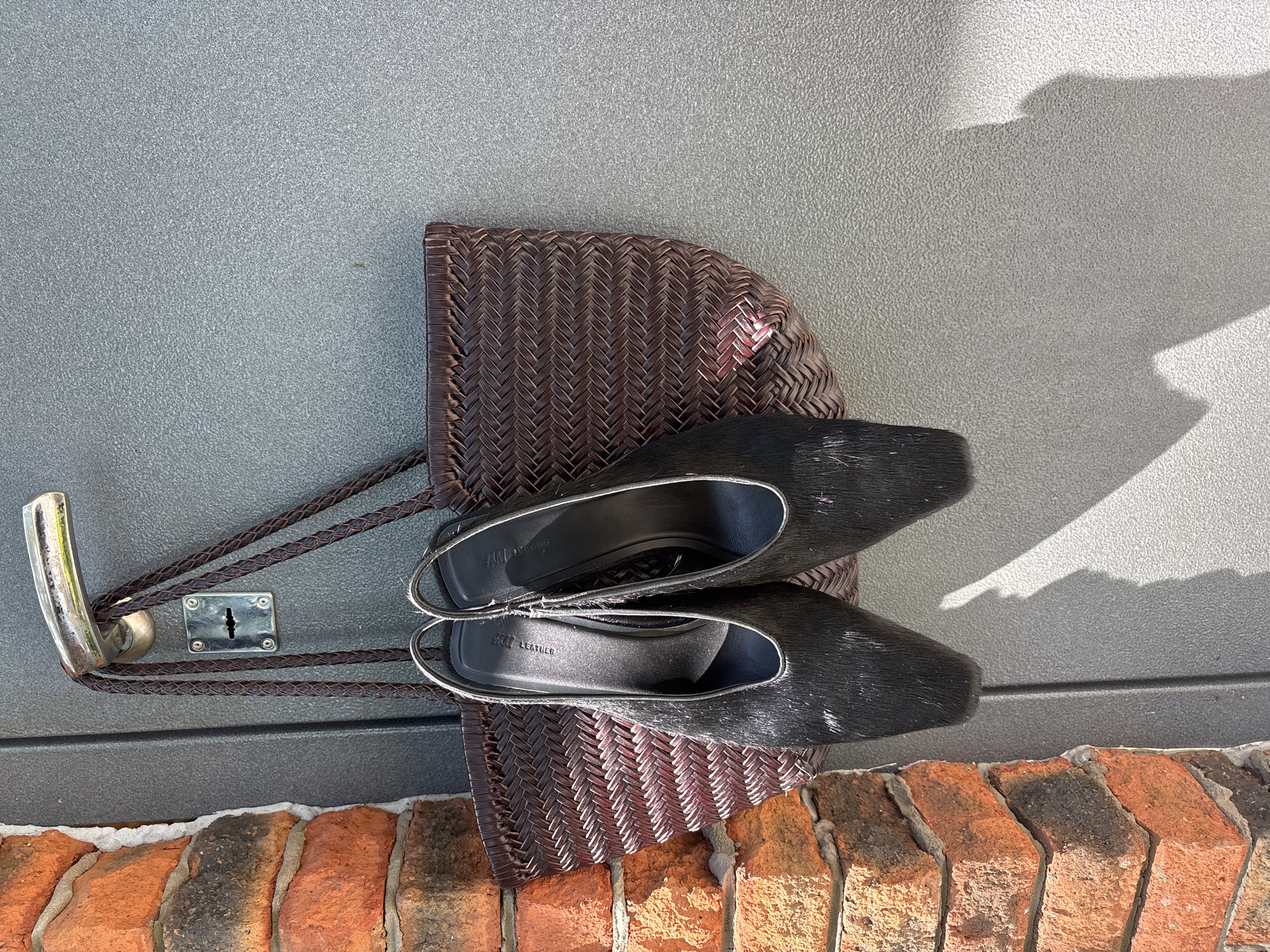 The image shows a brown woven bag hanging from a metal door handle of a gray door. Two black, fuzzy shoes rest on top of the bag. The bottom of the doorframe is brick. The location is Chaikovsky, Russia.
