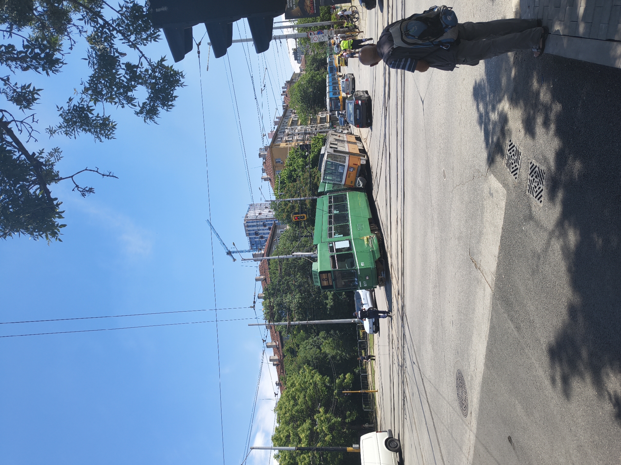 The image depicts an accident scene at a tram intersection in Sofia, Bulgaria. Two trams, one green and one orange and yellow, have derailed and are partially blocking the intersection. The green tram is largely upright, while the orange and yellow tram is leaning significantly.
Several people are present at the scene, including a person in a police uniform directing traffic and a person wearing a high-visibility vest, possibly a traffic controller or emergency responder. A man with a backpack stands on the sidewalk, observing the situation. In the background, a white van, a black car, and several other vehicles are visible. Buildings line the street, and trees provide shade. Power lines and traffic lights are present above the intersection. The sky is clear and blue.