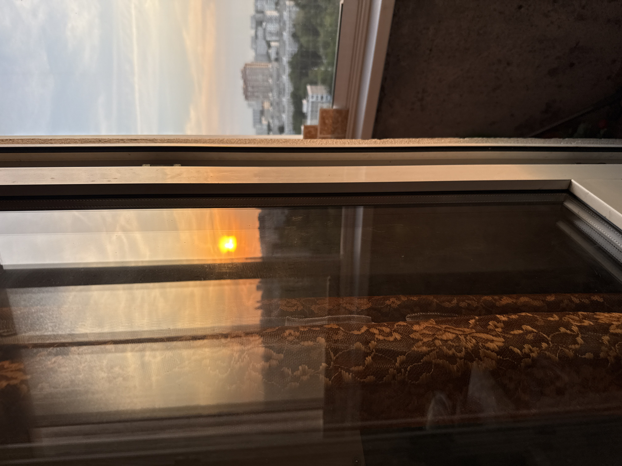 The image depicts an urban sunset viewed from inside a building in the city of Moscow, Russia, looking out through a white-framed window or balcony door. The left side of the frame shows the interior, where a dark orange-brown curtain with a distinct, intricate lace-like pattern is visible behind the glass. The glass itself reflects the warm, golden hues of the sunset and exhibits minor smudges and dust patterns.
The window frame, which appears to be a multi-pane structure, divides the view. To the right of the innermost white frame, the exterior is visible. A low, bright orange-yellow sun is prominent on the horizon, casting a strong glow and illuminating the sky, which transitions from vibrant orange near the sun to a pale blue-grey with faint cloud formations higher up.
Below the horizon, a sprawling cityscape extends into the distance, characterized by numerous multi-story buildings, primarily in white, grey, and light beige tones. Patches of green foliage from trees are interspersed between the structures. In the mid-ground, immediately outside the window frame to the right, a textured, rough-surfaced balcony wall or ledge is visible, along with a stack of light brown, rectangular objects, likely bricks or tiles, resting on a white windowsill or railing. No people are present in the scene.
