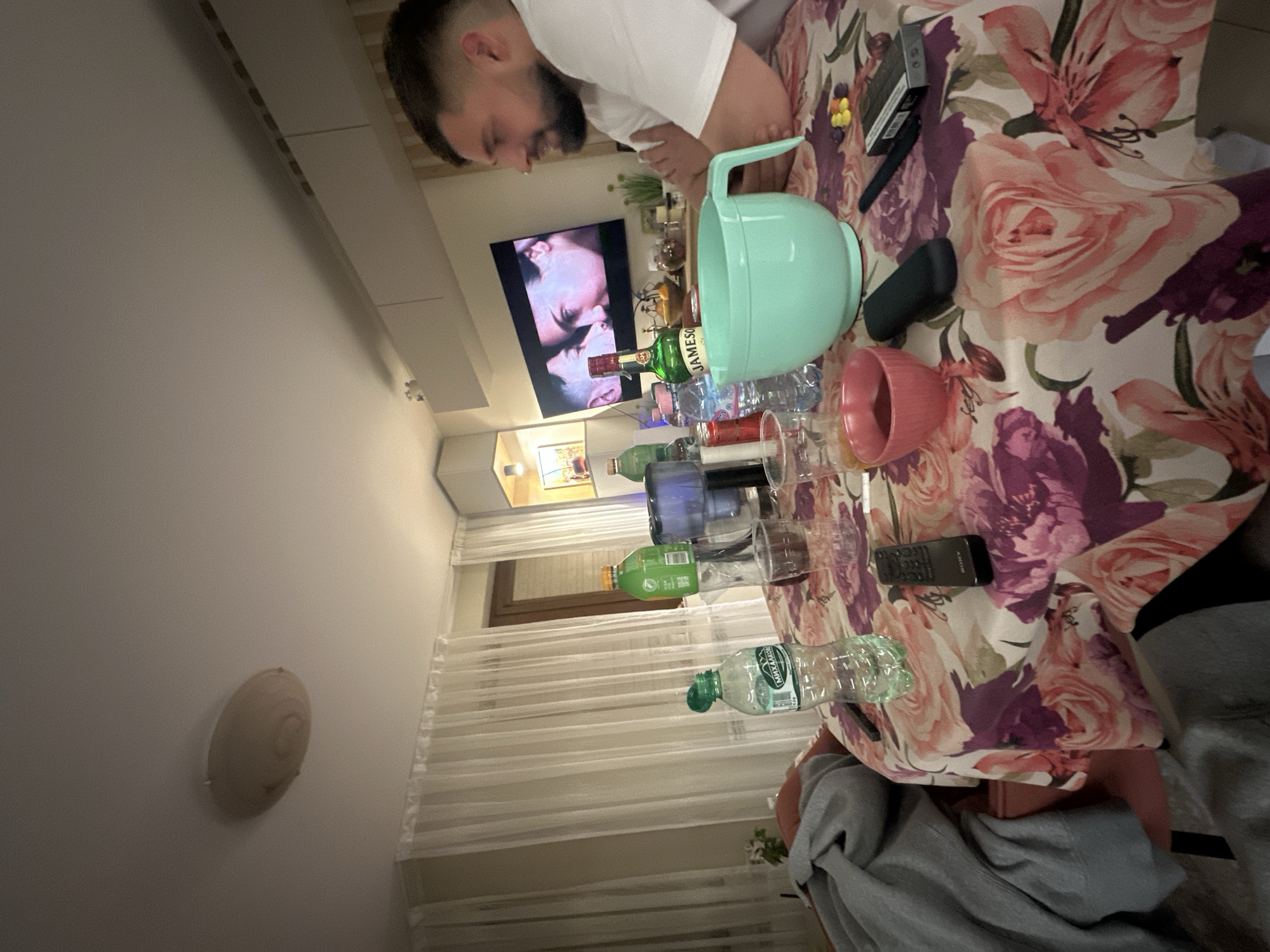A man with a beard wearing a white shirt is looking at a table covered with a floral tablecloth in an indoor setting. The table is cluttered with various items, including a green plastic mixing bowl, a bottle of Jameson Irish Whiskey, numerous water bottles, drinking glasses, a pink mug, a black rectangular device, and what appears to be a pack of cigarettes. In the background, a television is visible displaying a scene of two people embracing. The setting appears to be a residential room, with curtains and a wall-mounted light fixture visible. The location is unspecified but inferred by implication to be in Plovdiv, Bulgaria.