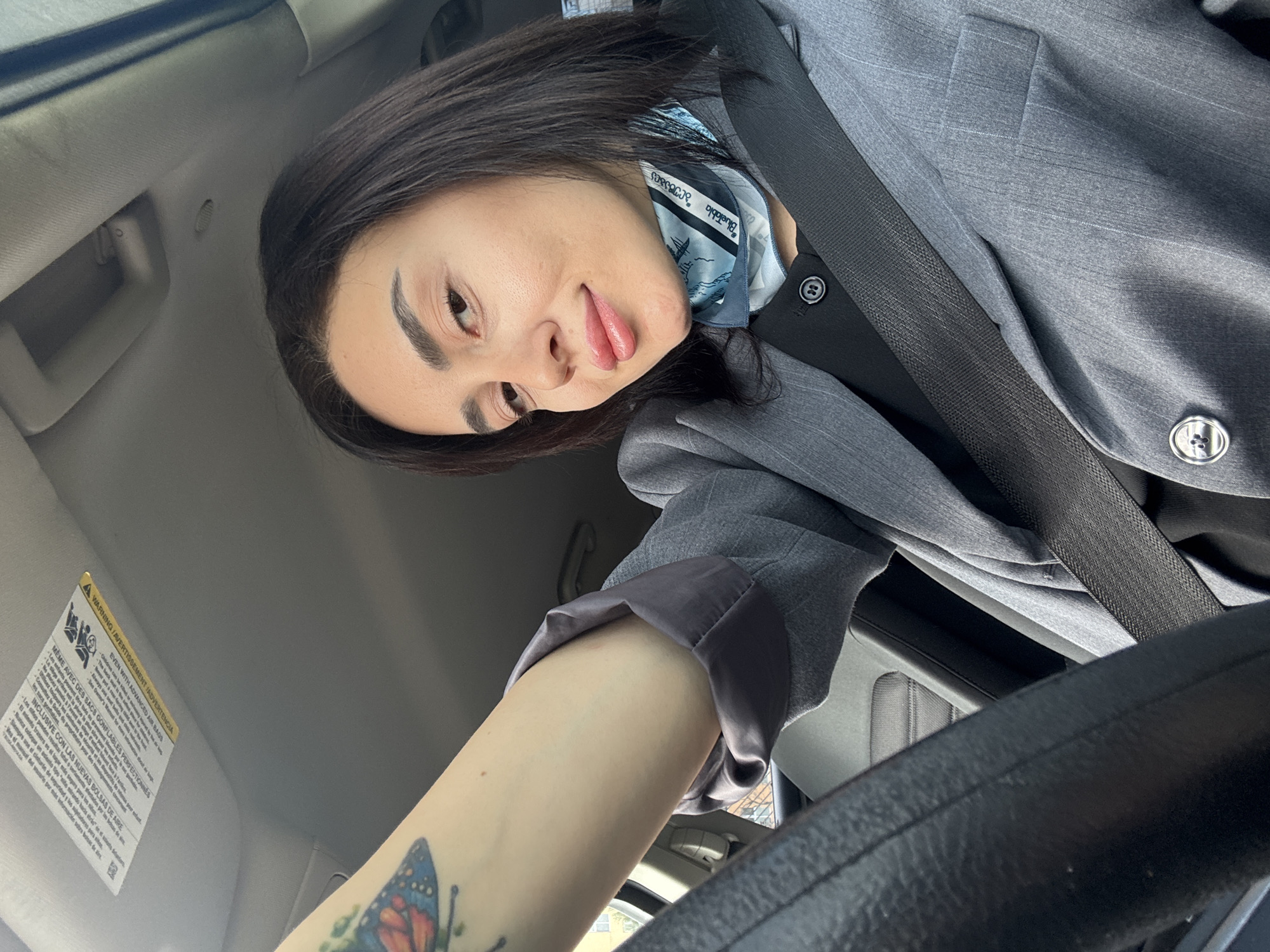 The image shows a person sitting in a car, wearing a gray blazer and a blue patterned scarf. They have a seatbelt on. The person has dark hair and appears to be smiling. A tattoo of a butterfly is visible on their arm. The car's interior, including the steering wheel and a warning label on the sun visor, is visible. The context suggests the person is in a car. The image does not provide any information about being in Tbilisi, Georgia.