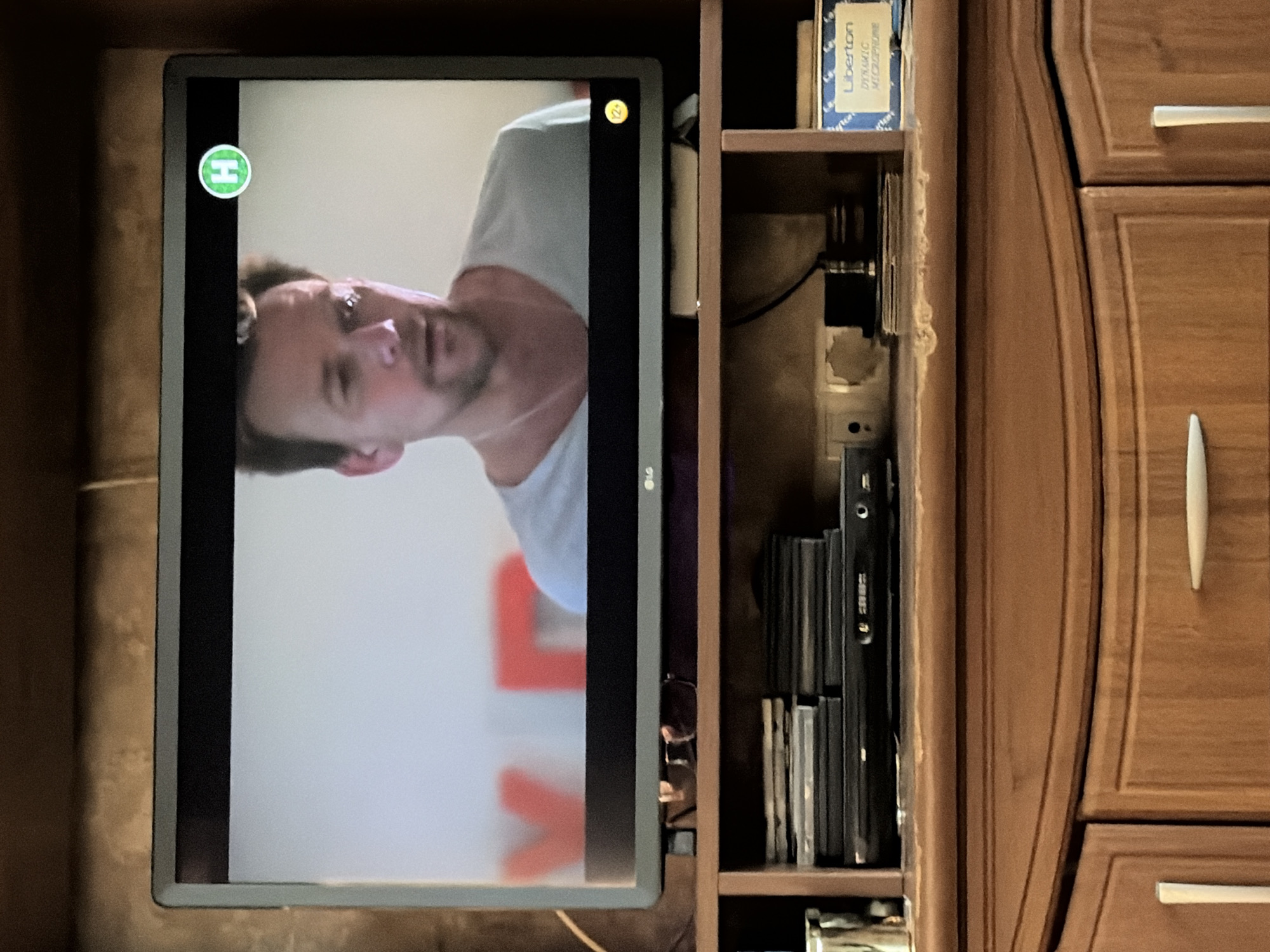 An overhead shot captures a television screen displaying a man with short brown hair and a light blue t-shirt. A green circular logo with an "H" is visible in the upper right corner of the screen, and a yellow circular logo with "12+" is in the lower right. The TV is placed on a wooden shelf unit, below which are several DVDs and a DVD player. To the right of the DVDs is a blue box labeled "Liberton Dynamic Microphone." The background of the image is a wooden wall.