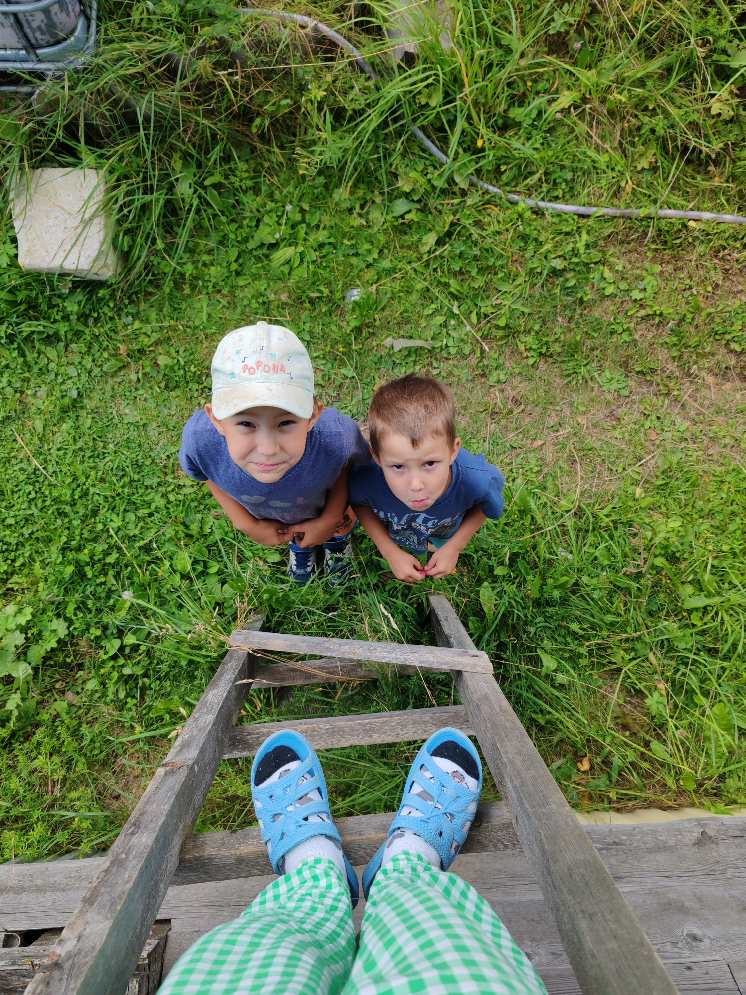 Two young boys stand on a wooden ladder, looking up at the camera. The boy on the left wears a light blue t-shirt and a white baseball cap with the word "POPOVA" written on it. He has a slight smile on his face. The boy on the right wears a dark blue t-shirt and has a neutral expression. Both boys are in a grassy area. The perspective of the photo suggests the camera is positioned from a higher vantage point, looking down on the boys. The environment appears to be outdoors, likely in a rural or suburban setting, given the presence of uncut grass and a simple wooden ladder. The image was captured in Chukhlem, Russia.