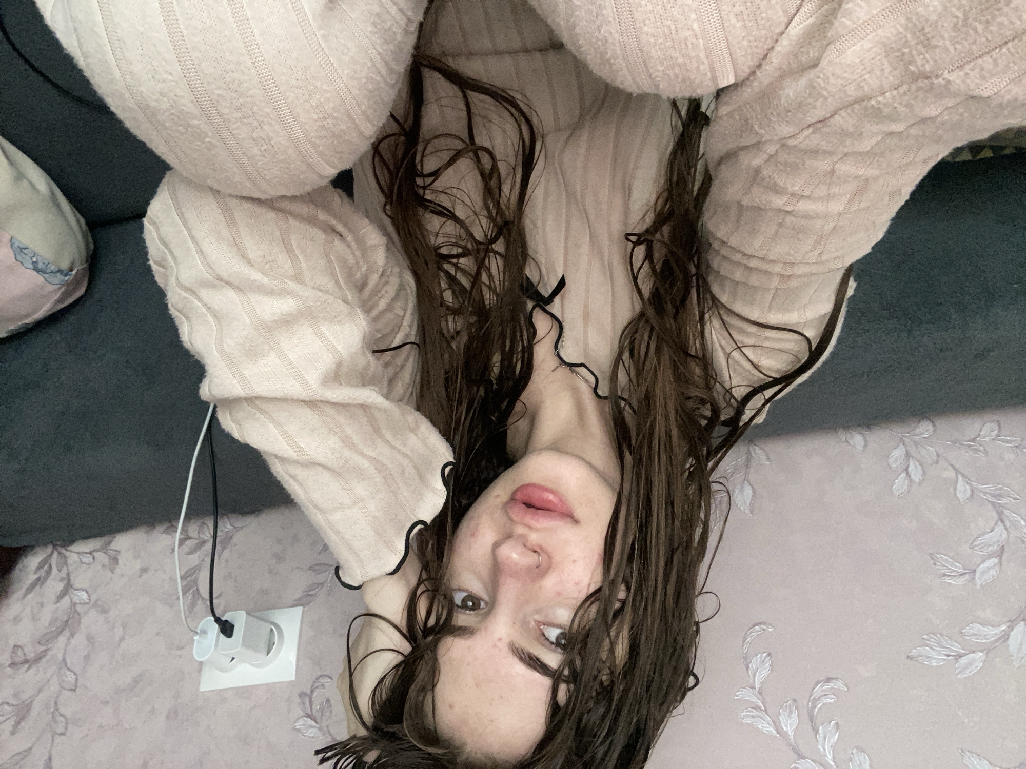 The image is a high-angle selfie of a young woman lying down indoors, taken from above and rotated 90 degrees clockwise. The woman's head is towards the bottom of the frame, and her upper body and arms are towards the top.
She has long, dark brown hair that appears wet or damp, styled to fall around her face and shoulders. Her gaze is directed straight at the camera with a neutral expression, lips slightly parted and possibly wearing lip gloss. A small metallic nose ring is visible in her left nostril. She is wearing a light pink or peach-colored ribbed sweater with a subtle black ruffled trim visible around the neckline. Her arms, covered by the sweater, are bent and positioned towards her head.
The background suggests an indoor setting. To the upper left, a dark grey or blue sofa or bed is partially visible, along with a patterned cushion in muted pink and blue tones. The right and bottom parts of the image show a light pink or lilac-colored wallpaper adorned with a delicate white, vine-like leaf pattern. A white wall socket is visible against the wallpaper, with a white charging adapter plugged in, from which a black cable and a separate white cable extend. The lighting is soft and even, indicating an indoor environment, possibly during the day or evening. No text is visible in the image.