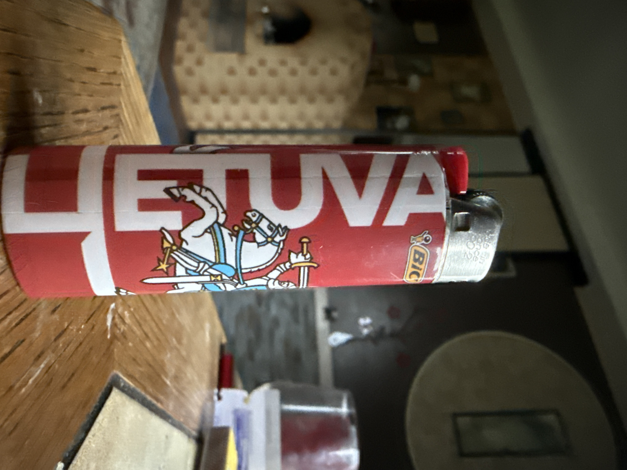 A close-up, slightly angled shot shows a red BIC lighter with white lettering that reads "LEVA" vertically down the side. The lighter also features a drawing of a knight on a white horse holding a sword. The lighter is standing upright on a wooden surface that appears to be a table. The background is blurred, but shows a wall with pictures and some circular decorative elements, possibly a tapestry or mural. To the left, a tan-colored, textured object with an arched opening is visible, resembling a bread oven or fireplace. The lighting is dim, suggesting an indoor setting, likely a home or a workshop. The scene is devoid of human presence, focusing solely on the object and its surroundings. There is no information available regarding the location in Galinė, Lithuania.