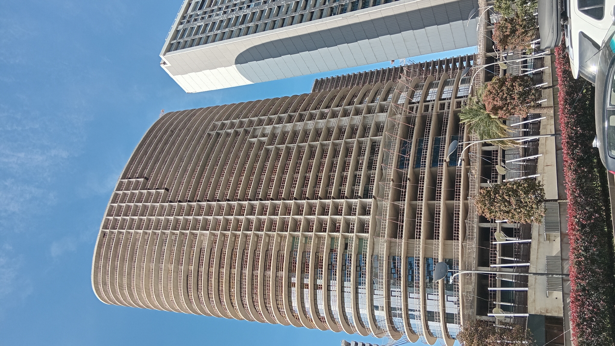 The image depicts a worm's-eye view of two buildings in Batumi, Georgia, under a clear blue sky. The foreground building is a curved, multi-story structure with a repeating pattern of vertical concrete ribs and horizontal beams, creating a grid-like façade. Scaffolding is visible on some sections, and some windows have been installed. The adjacent structure is a taller, rectangular building with a flat roof and a lighter-colored exterior. Street-level features include trees, bushes, street lamps, and parked cars along the right side of the frame. The photograph is taken from a low angle, emphasizing the height and architecture of the buildings.