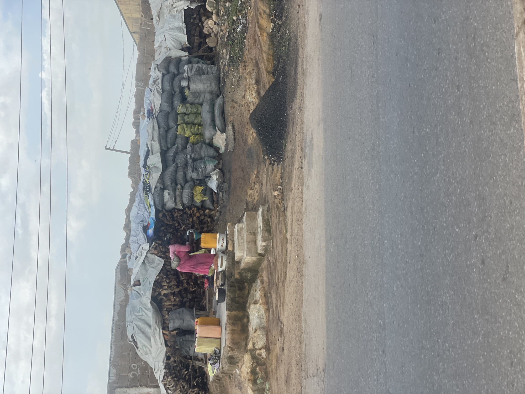 The image depicts a roadside market scene in Jos, Nigeria, with a focus on the sale of charcoal and firewood. In the foreground, a grey asphalt road is visible. Behind the road, a dusty, uneven ground slopes upwards towards a raised platform constructed from concrete and stone.
On this platform, a person wearing a pink garment is actively engaged in processing or preparing materials. Nearby, several large, colorful plastic containers (likely for water or other liquids) are placed, alongside bowls and what appear to be cooking vessels. A stack of firewood is visible to the left of the platform, covered in part by a white tarp.
Dominating the background is a large collection of bagged charcoal, neatly stacked and covered with tarps. Some bags are dark grey, while others are a pale yellow or green. More firewood is piled high behind the charcoal bags. A small, conical pile of loose charcoal is present on the ground in front of the bagged charcoal. The setting appears to be an open-air market or roadside stall, with a brick wall in the upper background and power lines overhead. The sky is overcast with clouds.