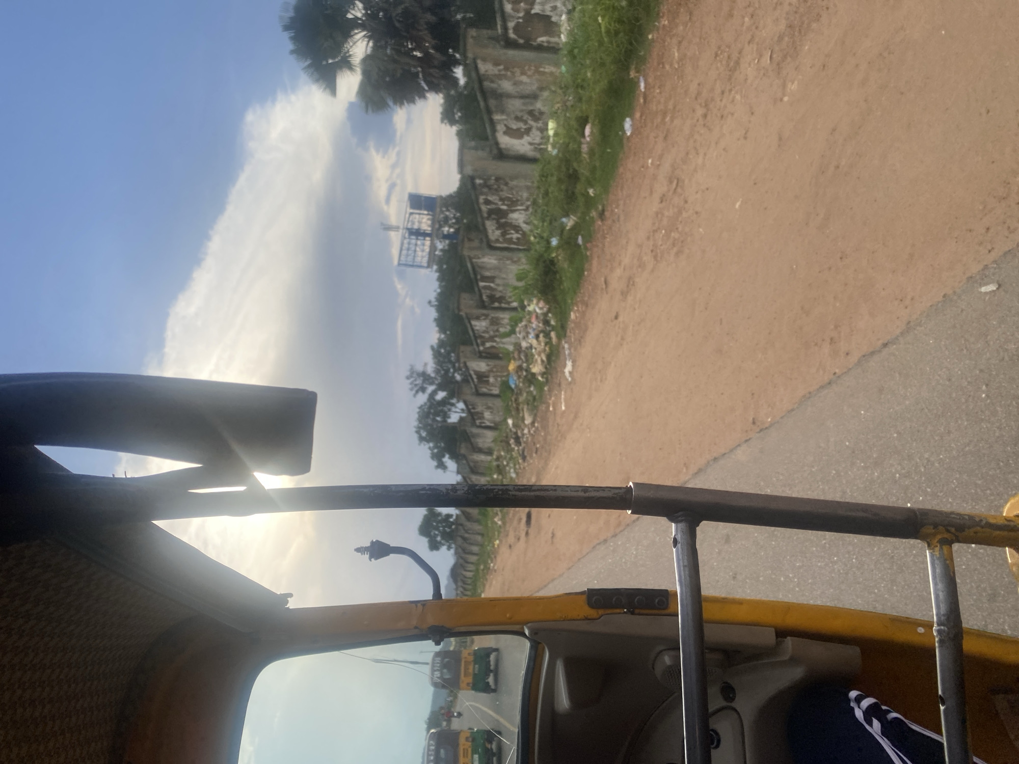 This image captures a perspective from inside a yellow auto-rickshaw, likely in Rafin Par, Nigeria. The view looks outwards towards a dusty, unpaved road on the right and a paved road on the left, where two other similar auto-rickshaws are visible in the distance. A long, weathered concrete wall runs along the right side of the road, lined with green foliage and debris, suggesting a roadside area. Above the wall, trees and a large billboard structure are discernible against a bright, cloudy sky. The sun is partially obscured by clouds and appears to be setting or rising, casting a bright light that creates some glare on the auto-rickshaw's interior. The interior of the auto-rickshaw shows the driver's side, including part of the dashboard, a seat, and metal bars forming the structure of the vehicle. The overall scene depicts a typical roadside environment in a developing urban or semi-urban area.