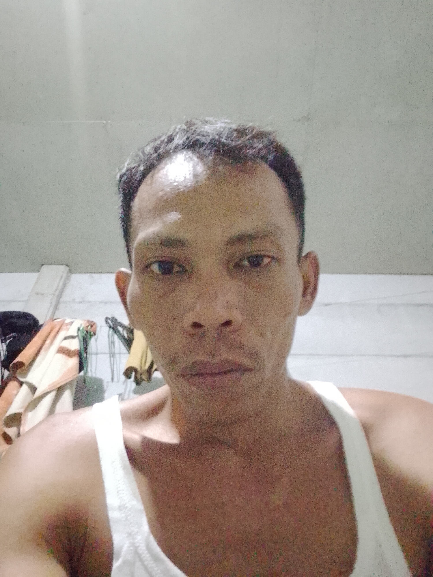 The image depicts a man in a white tank top, likely indoors, possibly in a residential setting. His hair is dark and short, with a receding hairline. The background features a light-colored wall and glimpses of what appears to be a cluttered area with hanging items, possibly clothes or towels. The location is specified as Bukit Ketok, Indonesia. No specific actions or interactions are evident in the frame.