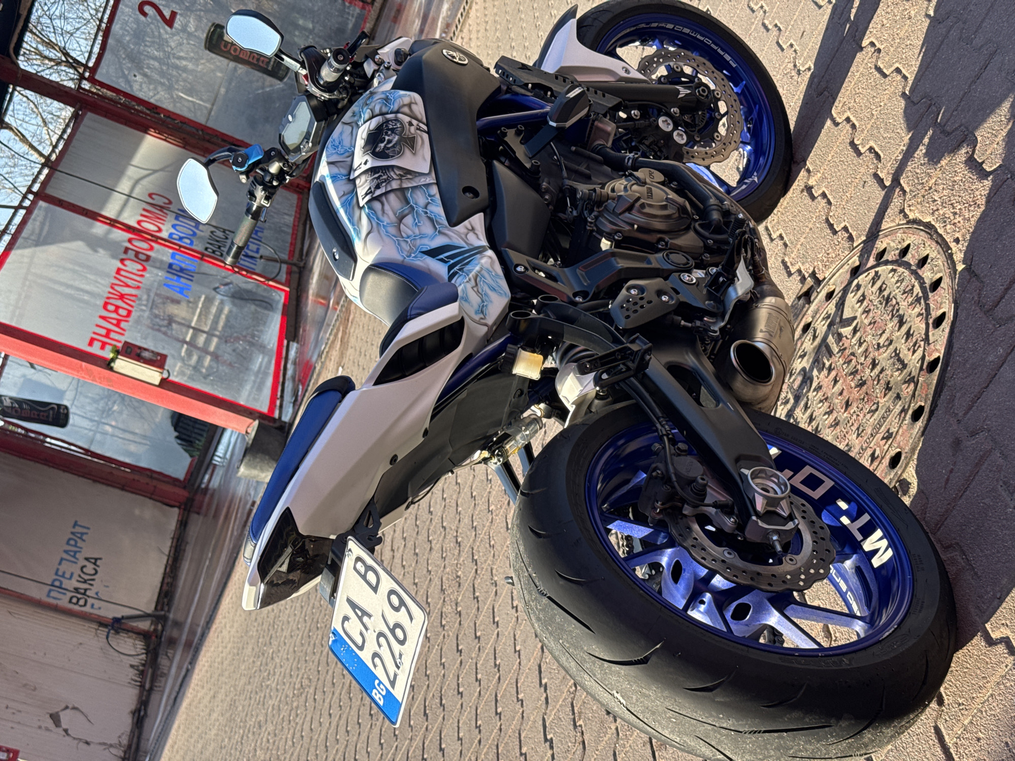 The image depicts a Yamaha MT-07 motorcycle parked on a paved surface. The motorcycle has a custom paint job with playing card and lightning bolt designs on the tank and side panels. The wheels are blue, with "MT-07" stickers on the front wheel. The license plate reads "CA B 2269 BG," indicating registration in Bulgaria. The motorcycle is parked next to a car wash facility with signage in Bulgarian. The overall scene suggests a sunny day. Based on the Bulgarian license plate and signage, it is highly probable that the location is in Bulgaria, and Sofia is its capital.