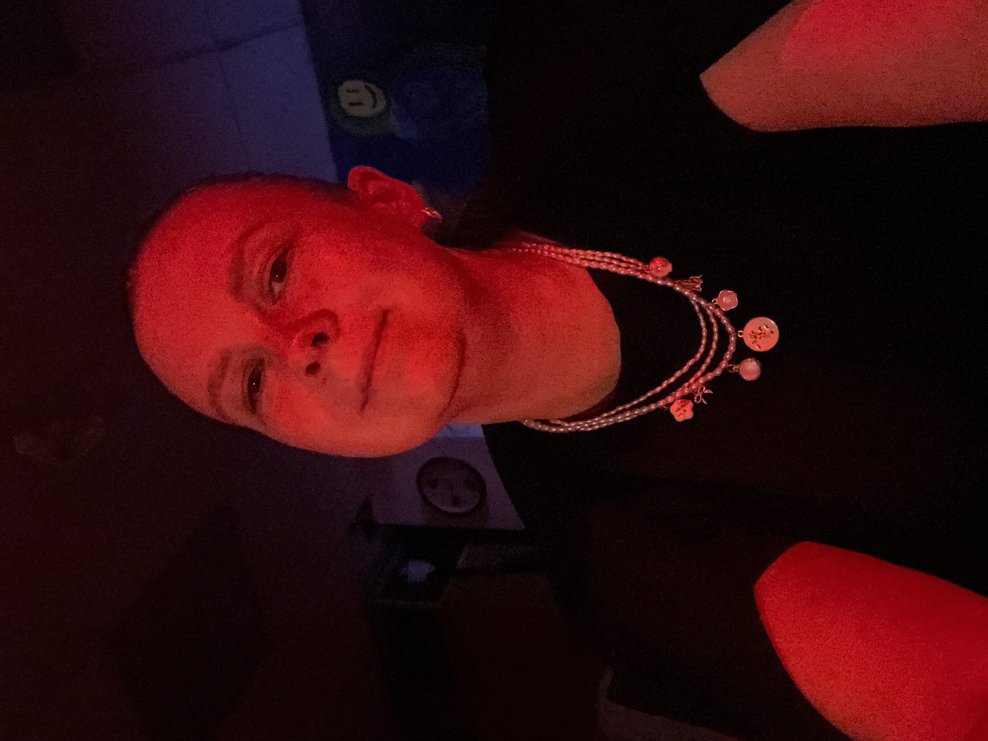 The image captures a person, likely a young woman or girl, illuminated by a dominant red light source, casting a strong crimson glow across her face and right arm. She is looking towards the camera with a neutral expression, possibly a subtle smirk. Her dark hair is pulled back, and she is wearing a dark-colored top or dress.
Around her neck, she accessorizes with a multi-strand necklace, appearing to be made of small, light-colored beads. This necklace is adorned with several charms, including a prominent circular disc with an engraved design, and smaller charms shaped like flowers or hearts.
The background is dimly lit and dark, indicating an indoor setting, likely at night or in a room with specialized lighting. In the upper right quadrant of the background, a dark surface, possibly a wall or furniture, displays a bright green object with a distinctive smiley face symbol. Further down and to the left, another circular object, potentially a small clock or a decorative element, is faintly visible on a wall. The artificial red light creates a specific mood. No legible text is visible in the image. The scene is consistent with an indoor environment, potentially in Chaikovsky, Russia.