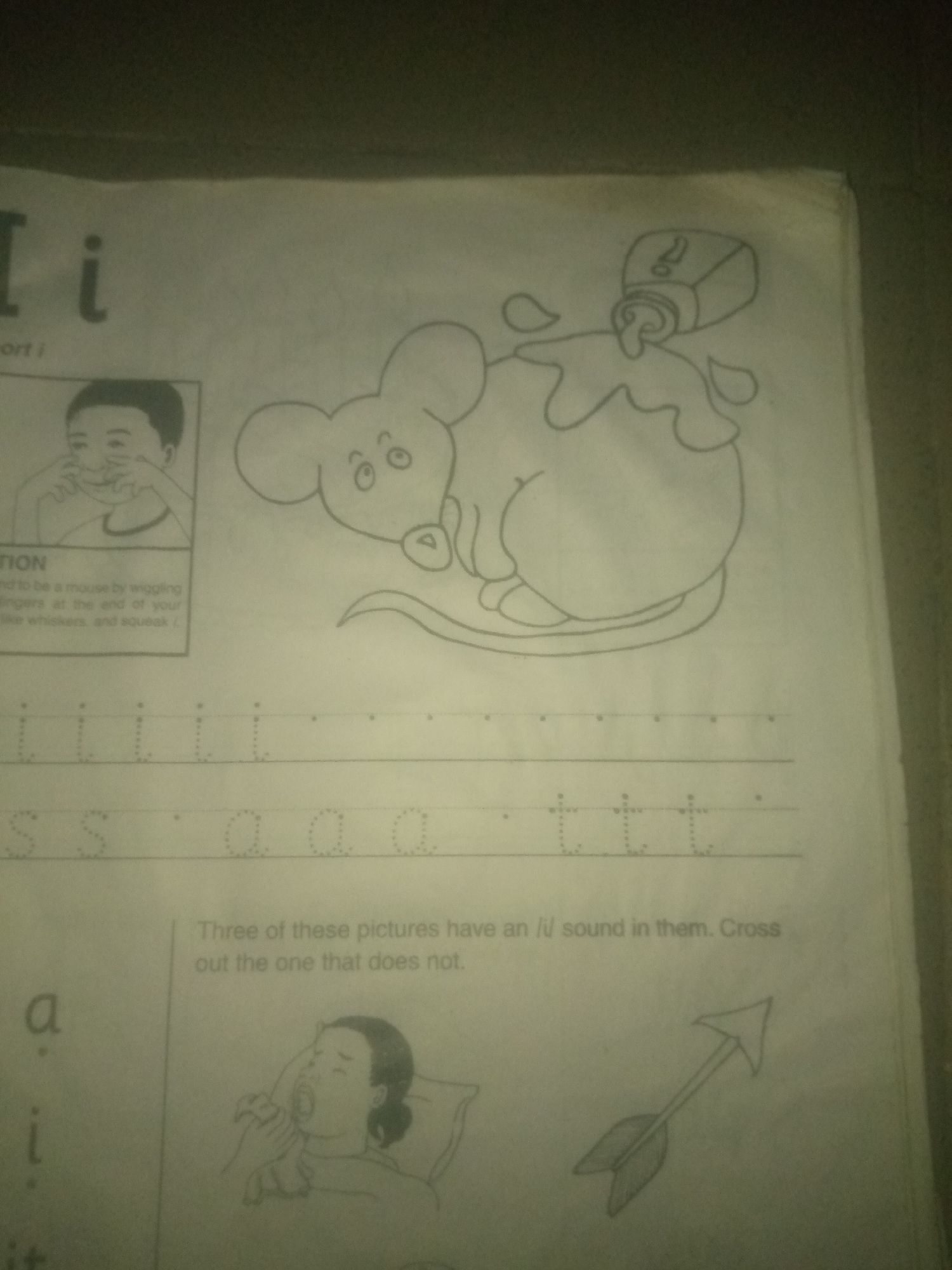 The image is a page from a children's learning worksheet. At the top left, the letters "I i" are printed in large font. Below this, a small box contains an illustration of a boy making a funny face, and some text that is partially obscured. To the right of the "I i" is a large drawing of a mouse with a bottle of liquid pouring onto it, creating a puddle. Below the mouse and the boy's illustration, there are lines with dotted letters for tracing: "i i i i i", "s s a a a", and "t t t". Further down, there is text that reads, "Three of these pictures have an /i/ sound in them. Cross out the one that does not." Below this text, on the left, are the letters "a" and "i" printed. To the right of these letters, there is an illustration of a person in bed, holding their throat and appearing to be sick. To the right of that is a drawing of an arrow. The worksheet appears to be old, with some yellowing and wear on the paper.
