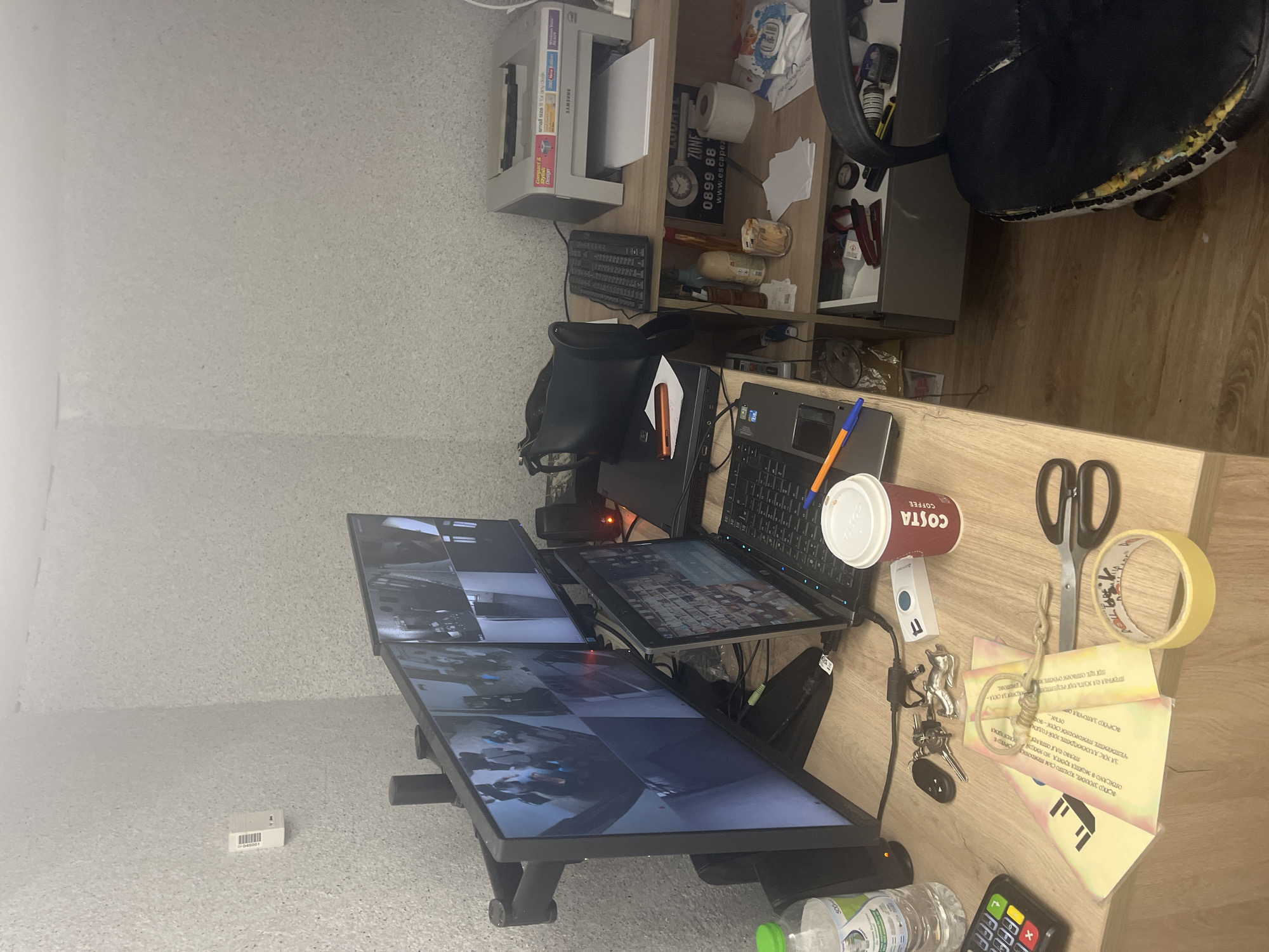 The image shows a cluttered desk area. In the foreground, there is a wooden desk surface with various items, including a calculator, keys, scissors, a roll of tape, a white paper cup with "COSTA COFFEE" printed on it, and some documents. There is also a laptop open on the desk. Behind the desk, there are multiple computer monitors displaying what appear to be security camera feeds. To the right of the monitors, there is a shelf unit with a printer on top, and various items on the shelves below, including bottles, a glass jar with money, and a roll of toilet paper. A black office chair with damaged upholstery is visible in the foreground on the right. The walls appear to be textured and light-colored.