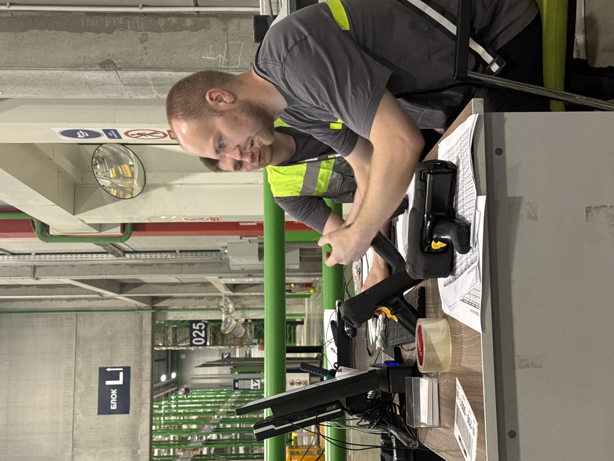 In Lupanovo, Russia, two men are seated at a desk in what appears to be a warehouse or industrial setting. The man in the foreground, wearing a grey t-shirt, is looking towards the left and gesturing with his hands. He has a short, light brown haircut and is facing a computer monitor. To his left, a second man, wearing a high-visibility vest over a t-shirt, is also looking towards the left, seemingly engaged in the same task or conversation. The desk is cluttered with various items including computer equipment, a scanner, a roll of tape, papers, and a small box of cards. In the background, rows of green shelving units are visible, typical of a warehouse, with signage indicating "БЛОК L" and "025". The environment suggests a functional workspace within a large storage facility.