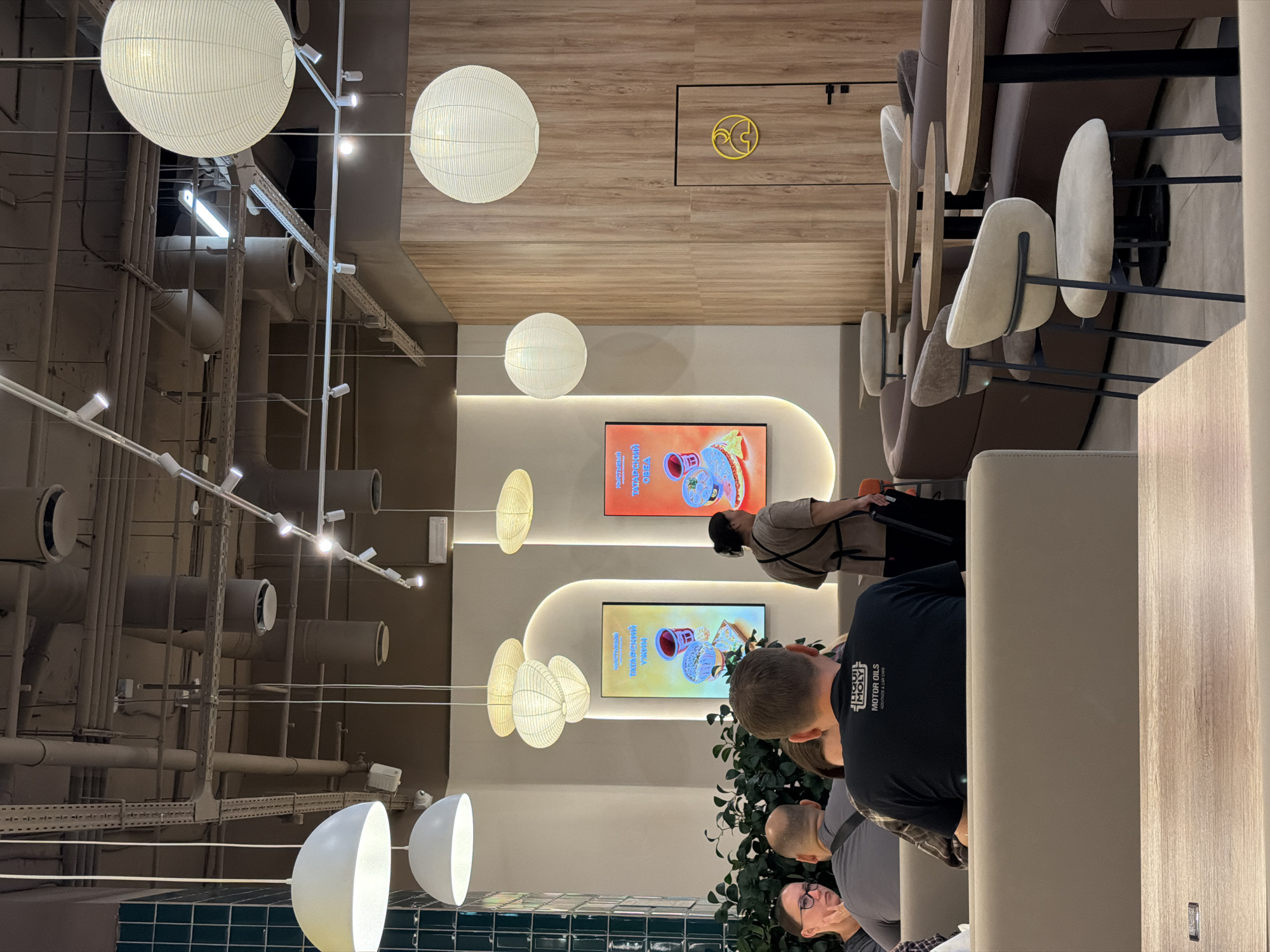 The image depicts the interior of a modern, casual dining establishment. Several large, white, spherical paper lanterns hang from the ceiling, providing ambient lighting. In the foreground, a group of diners are seated at light-colored booth-style seating. The focus is on the back of a man wearing a black t-shirt with white text. To his left, a woman with glasses is visible. In the mid-ground, a server with dark hair, wearing a light brown apron over a tan shirt, stands near a display board with images of food. Two digital menu boards, one with a yellow and blue theme and another with an orange and pink theme, are mounted on a curved wall. The walls are a mix of light beige and warm-toned wood paneling. Further back, round tables with dark bases and wooden tops are arranged with chairs, including some upholstered in light beige. The overall impression is a clean, contemporary space designed for casual dining.
