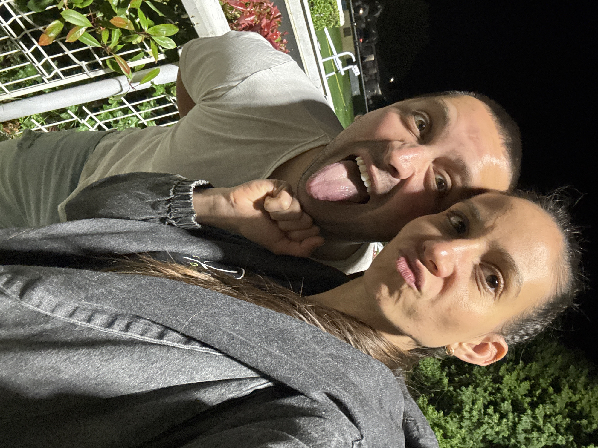 A man and woman are posing for a selfie outdoors at night in the Borough of Spelthorne, United Kingdom. The man, who is behind the woman, is wearing a light grey t-shirt and sticking his tongue out. The woman is wearing a grey jacket. There are plants and a metal fence visible in the background.