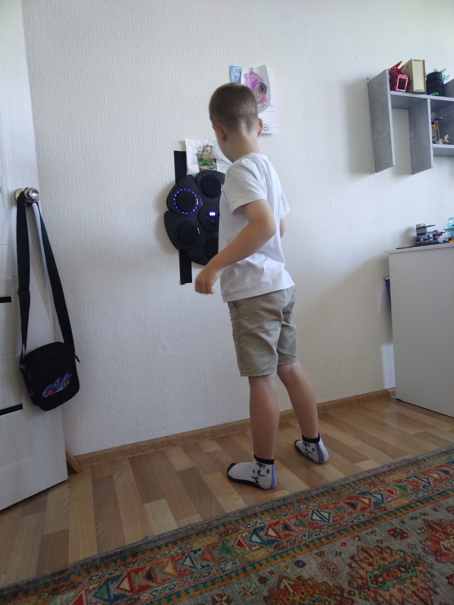 A young boy wearing a white t-shirt and khaki shorts stands in front of a black, circular device mounted on a textured white wall. The device has several illuminated purple circles and a small digital display. To the left of the device, a black messenger bag with a car graphic is hanging on a hook attached to a white door frame. On the wall above the device, there are several colorful drawings. To the right of the boy, a grey, multi-tiered shelf is mounted on the wall, containing various items including toys and books. In the foreground, a wooden floor is partially covered by a patterned rug. The lighting appears to be natural, coming from an unseen window. The overall impression is an indoor setting, likely a child's room or play area, in Богандинский, Russia.
