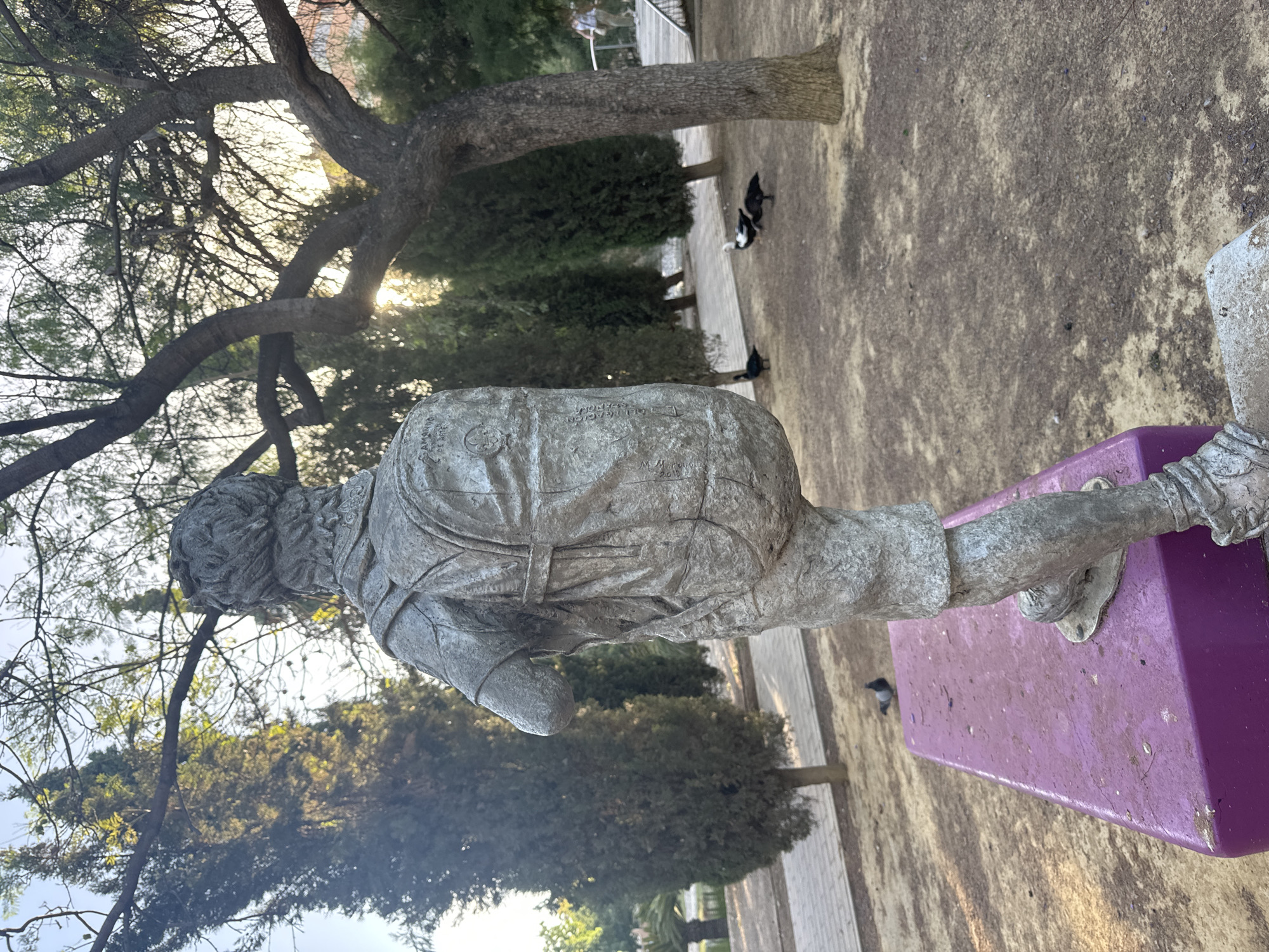 The photograph captures a statue of a young boy from behind, wearing a backpack and shorts, positioned on a purple pedestal in a park. The statue appears to be made of metal and is weathered. Several ducks are visible in the background on the ground, and a pigeon is perched on the pedestal near the statue's feet. The park setting features trees, a paved walkway, and a grassy area with scattered dry leaves. In the far background, a person is walking on an elevated pathway. The overall lighting suggests it is daytime, possibly late afternoon, with sunlight filtering through the trees. The presence of the statue, ducks, and trees indicates an outdoor public space. The location is identified as the 'Sculpture Garden of Famine and Children' in the city of Murcia, Spain.