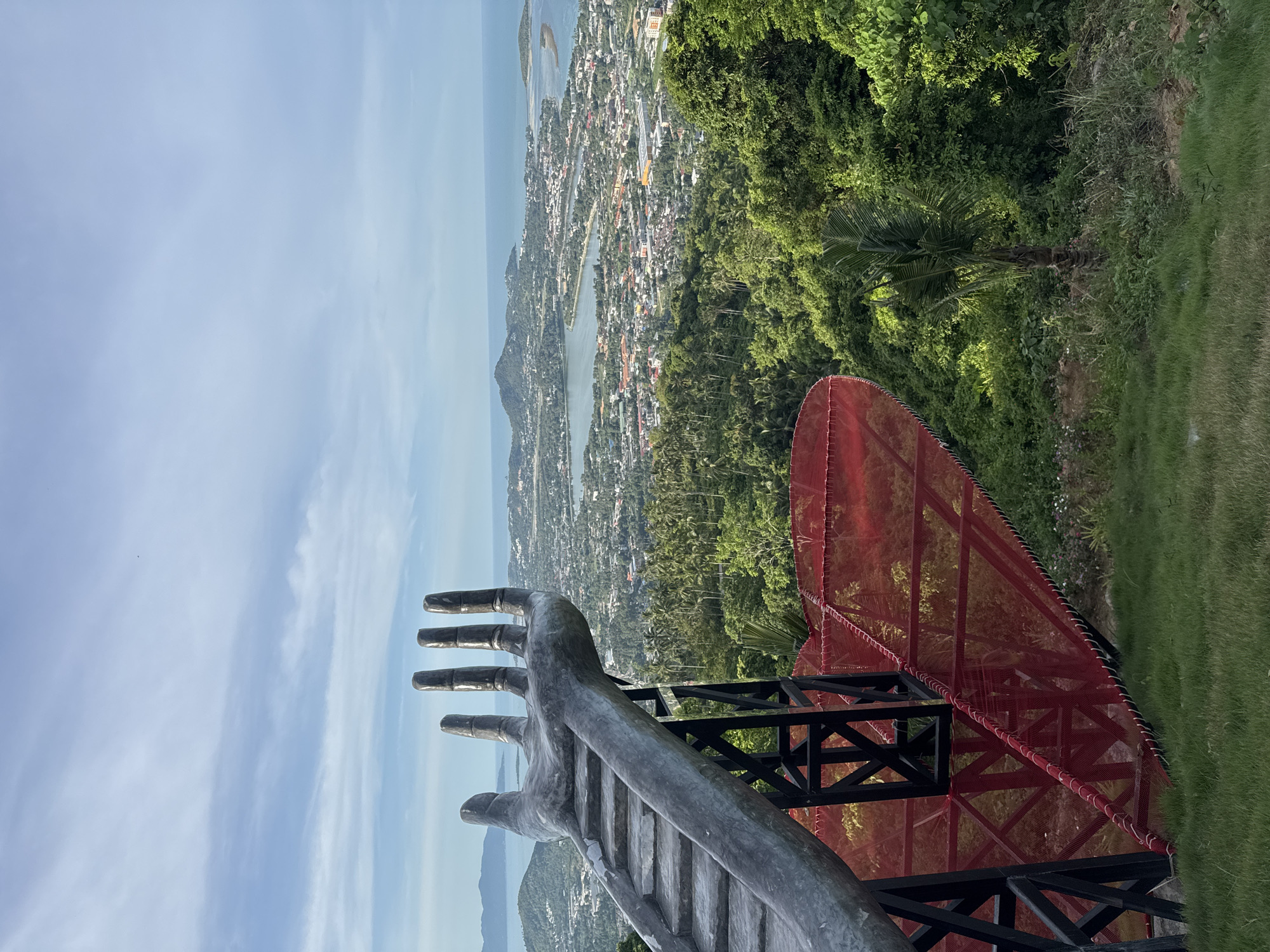 A hand-shaped sculpture with stairs leading up to it is in the foreground, overlooking a city and a bay. The sculpture is made of metal and has a weathered appearance. The stairs are concrete and appear to be worn. A red net structure is below the sculpture, which is supported by black metal beams. The city is nestled among lush green hills and palm trees, with a body of water in the background. The sky is partly cloudy with a few wispy clouds. The overall scene is scenic and evokes a sense of elevation and perspective.