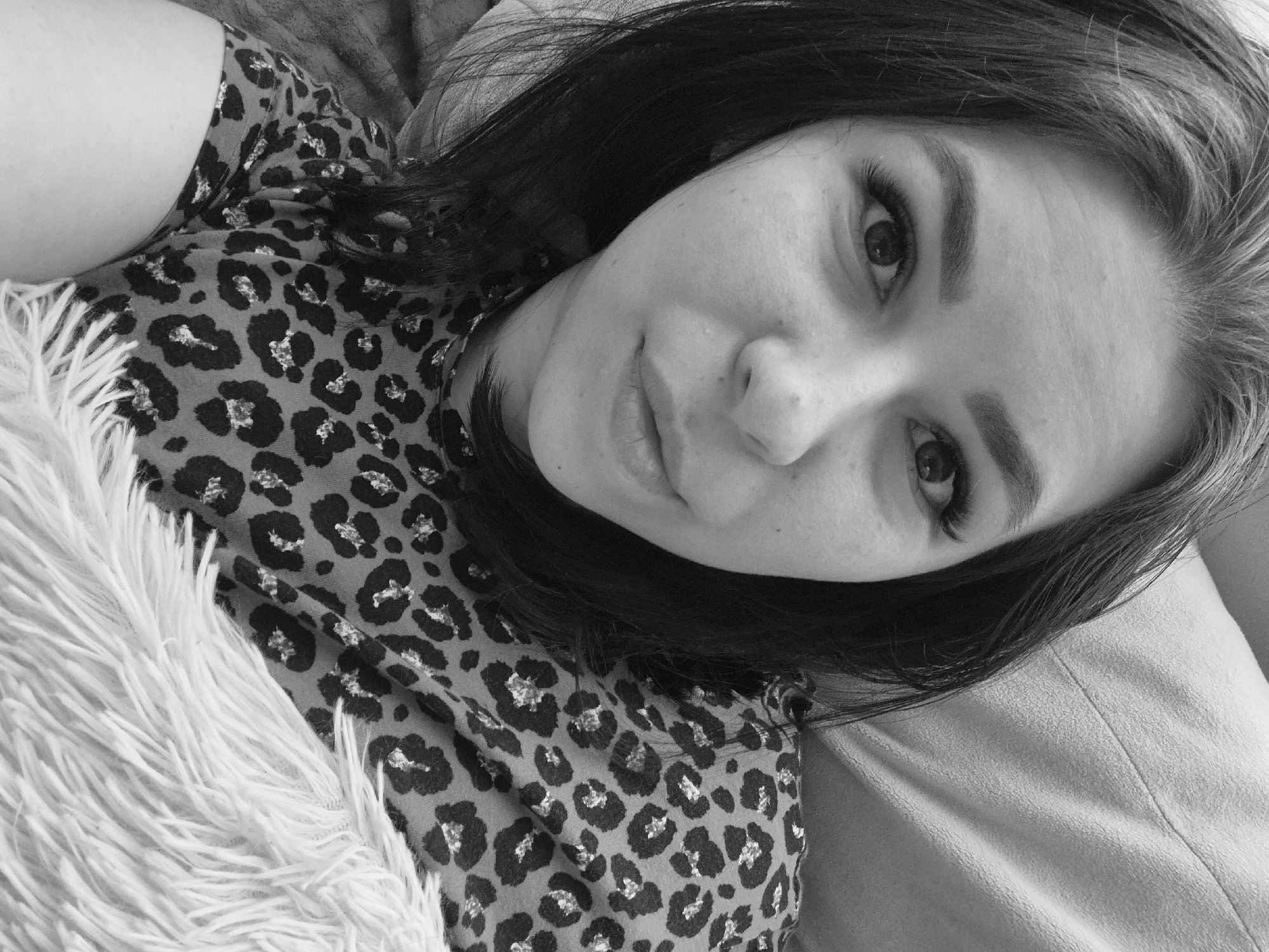 A grayscale, close-up, high-angle shot depicts a person with dark hair looking up towards the camera. The person is wearing a leopard-print garment. A fluffy white object appears on the left side of the frame, and a lighter-colored, solid material is visible on the right. No details indicate the location of Tobolsk, Russia.