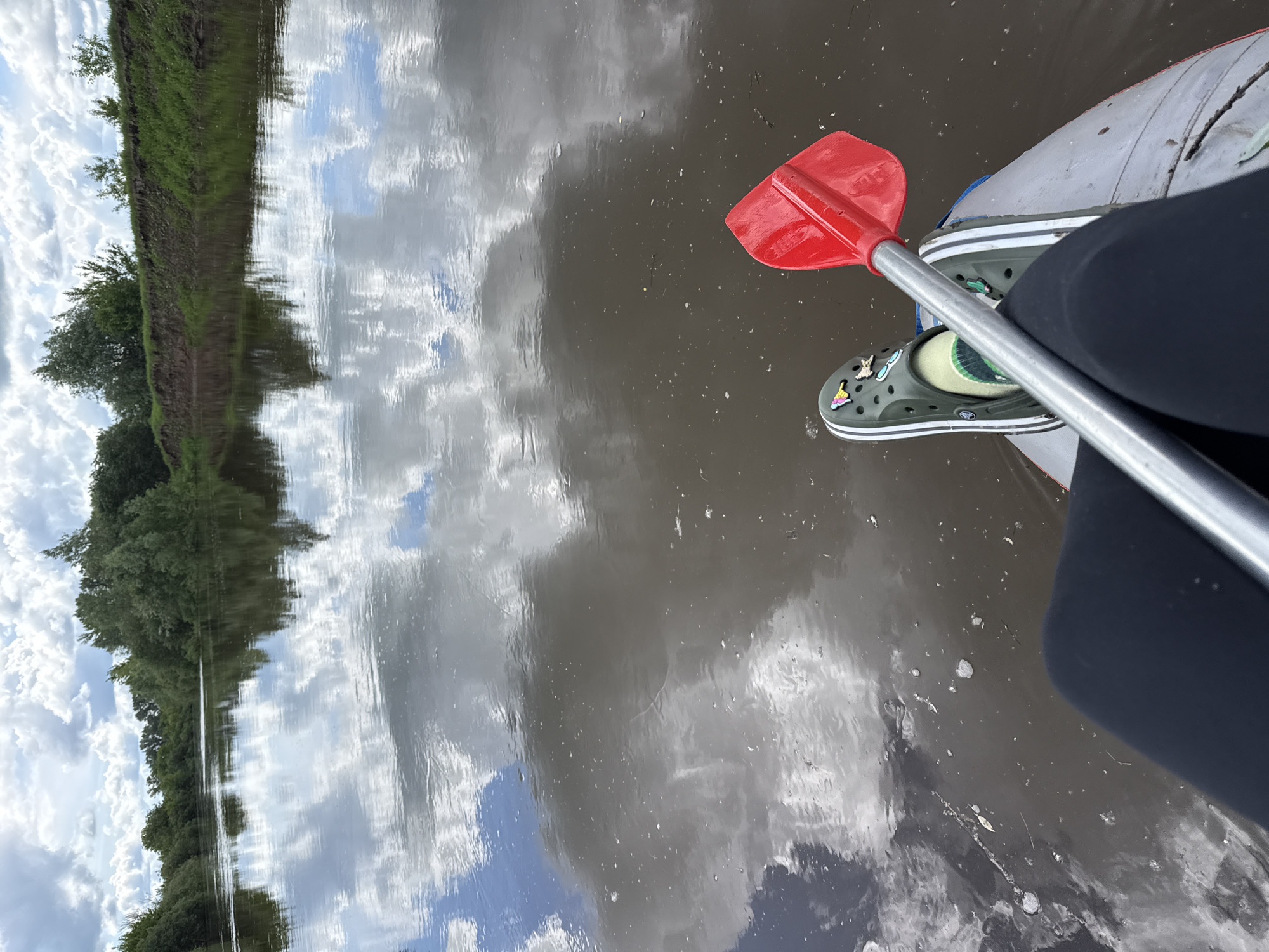 The image shows a person kayaking on a body of water in Izh-Boyki, Russia. The kayak is grey and white, with a person wearing black leggings and green Crocs inside. The kayak paddle is red and silver and lies across the person's lap. The water reflects the sky, which is filled with clouds. Trees and greenery line the water's edge on one side.
