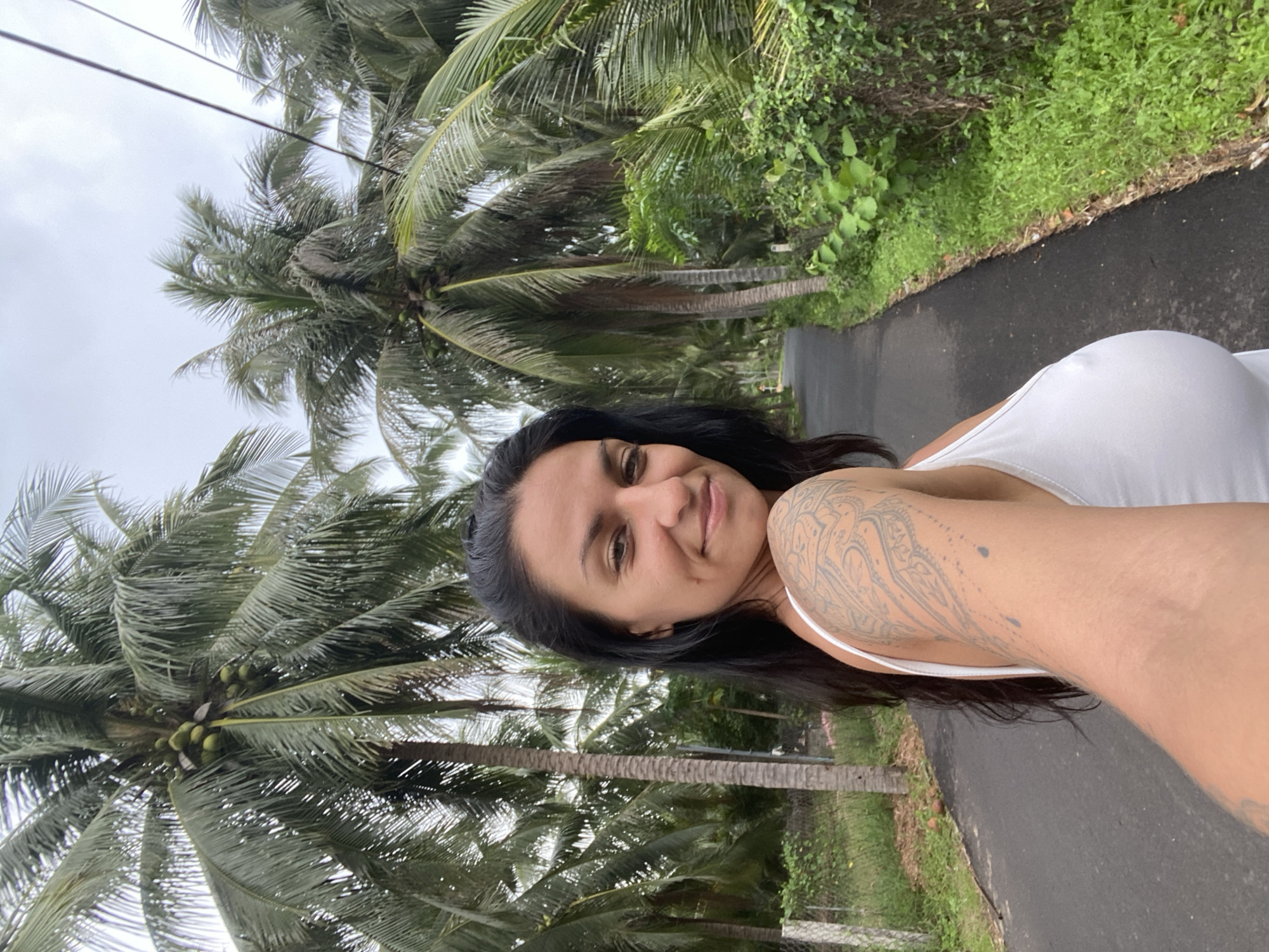 A woman with dark hair and a tattoo on her arm takes a selfie on a paved road lined with palm trees. The woman is wearing a white tank top. The road curves into the distance, and the sky is overcast. The palm trees are dense, and some have coconuts. The scene appears to be in Mũi Né, Vietnam, a coastal resort town known for its beaches and sand dunes.